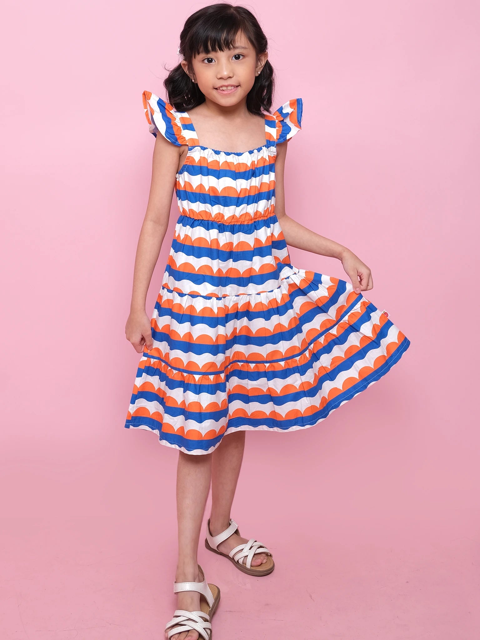 A young girl models the Nautical Holiday Dress by Nautical, featuring orange, blue, and white patterns, as she poses against a pink background and holds out part of her skirt.