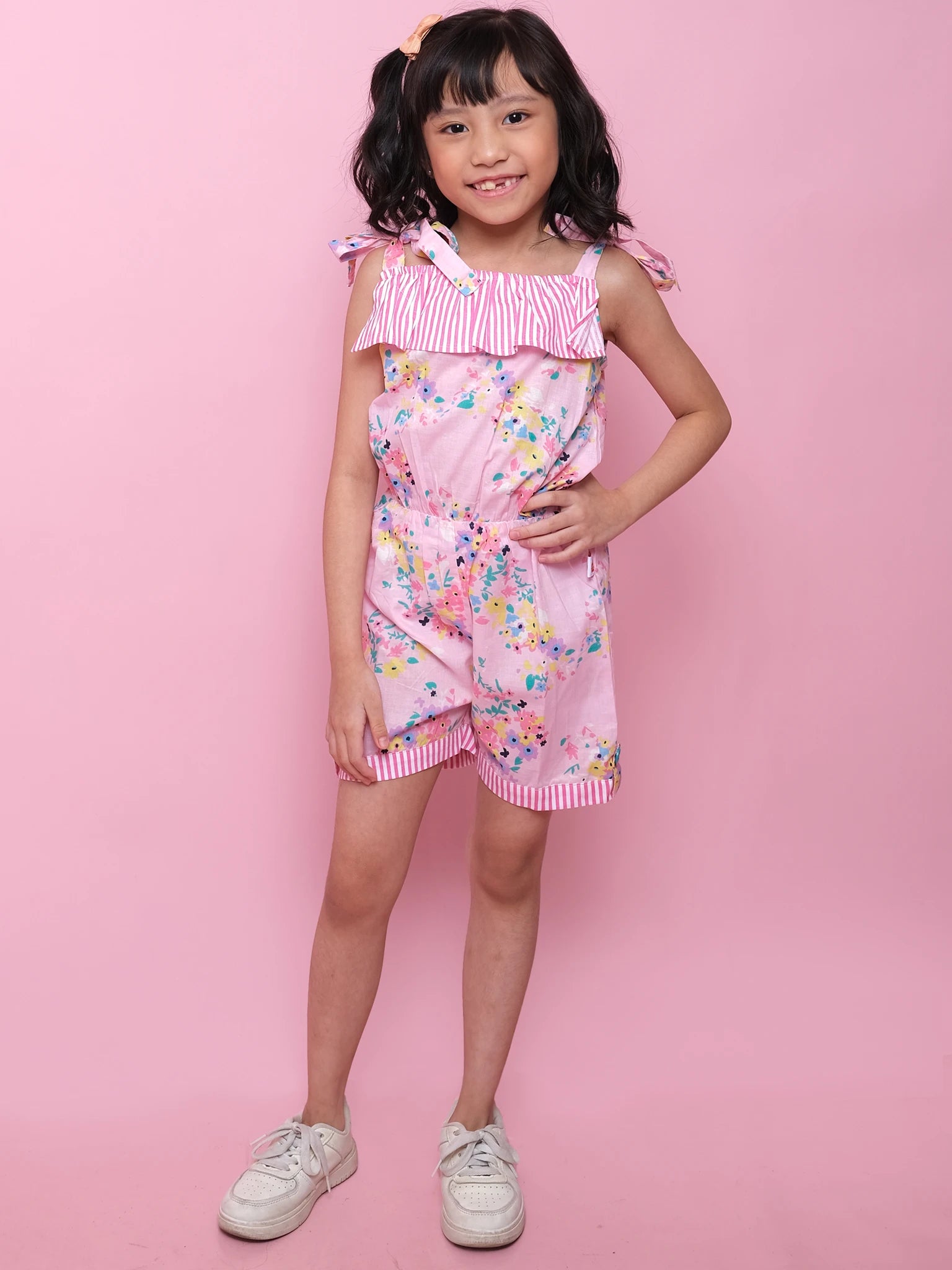 A young girl smiles at the camera in the Confetti Floral Josie Playsuit by Confetti Floral—a stylish pink floral cotton playsuit with striped details, paired with white sneakers—for a perfect summer look against a pink background.