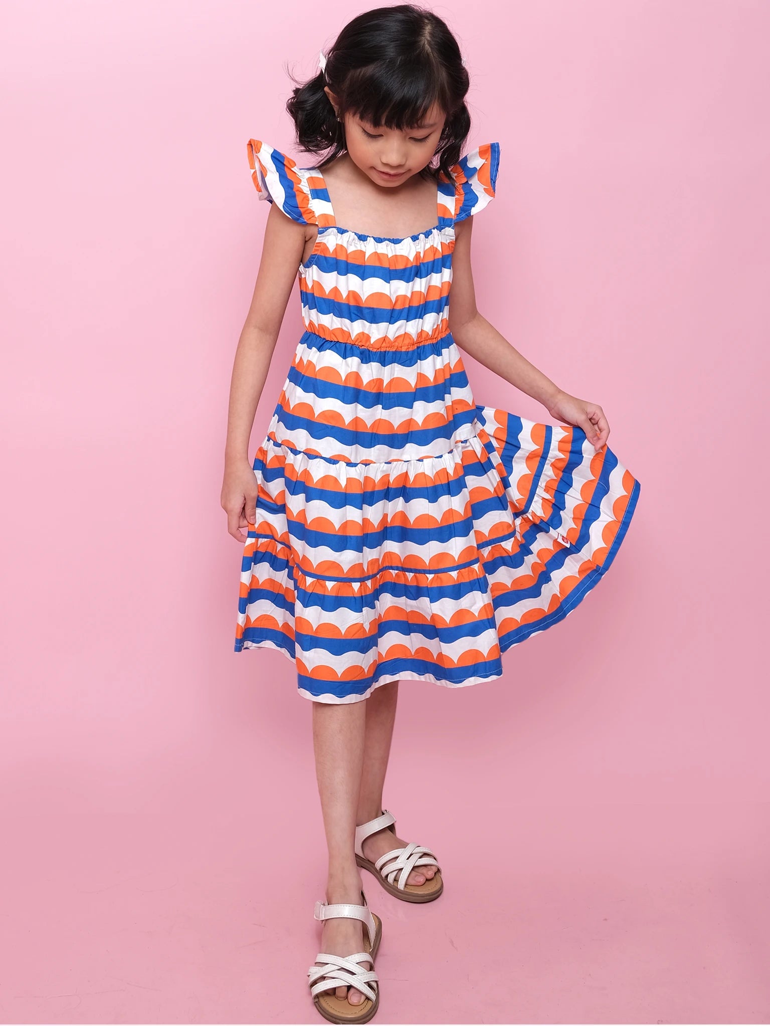 A young girl in the Nautical Holiday Dress by Nautical, featuring colorful stripes and ruffled sleeves, stands against a pink background, holding her skirt and looking down. She completes the look with white sandals.