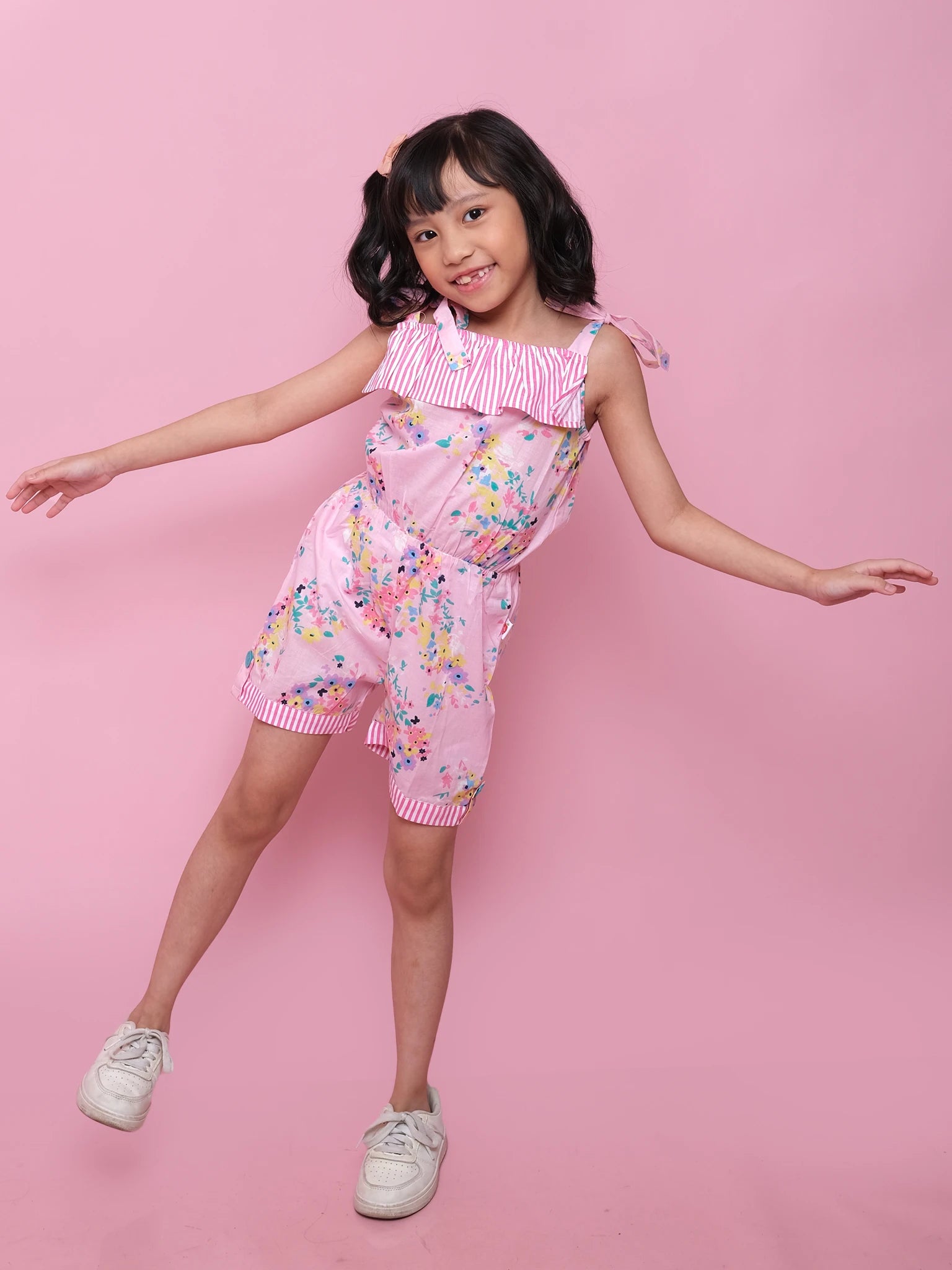 A young girl poses playfully against a pink background in the Confetti Floral Josie Playsuit by Confetti Floral, a cotton outfit ideal for fun summer style.