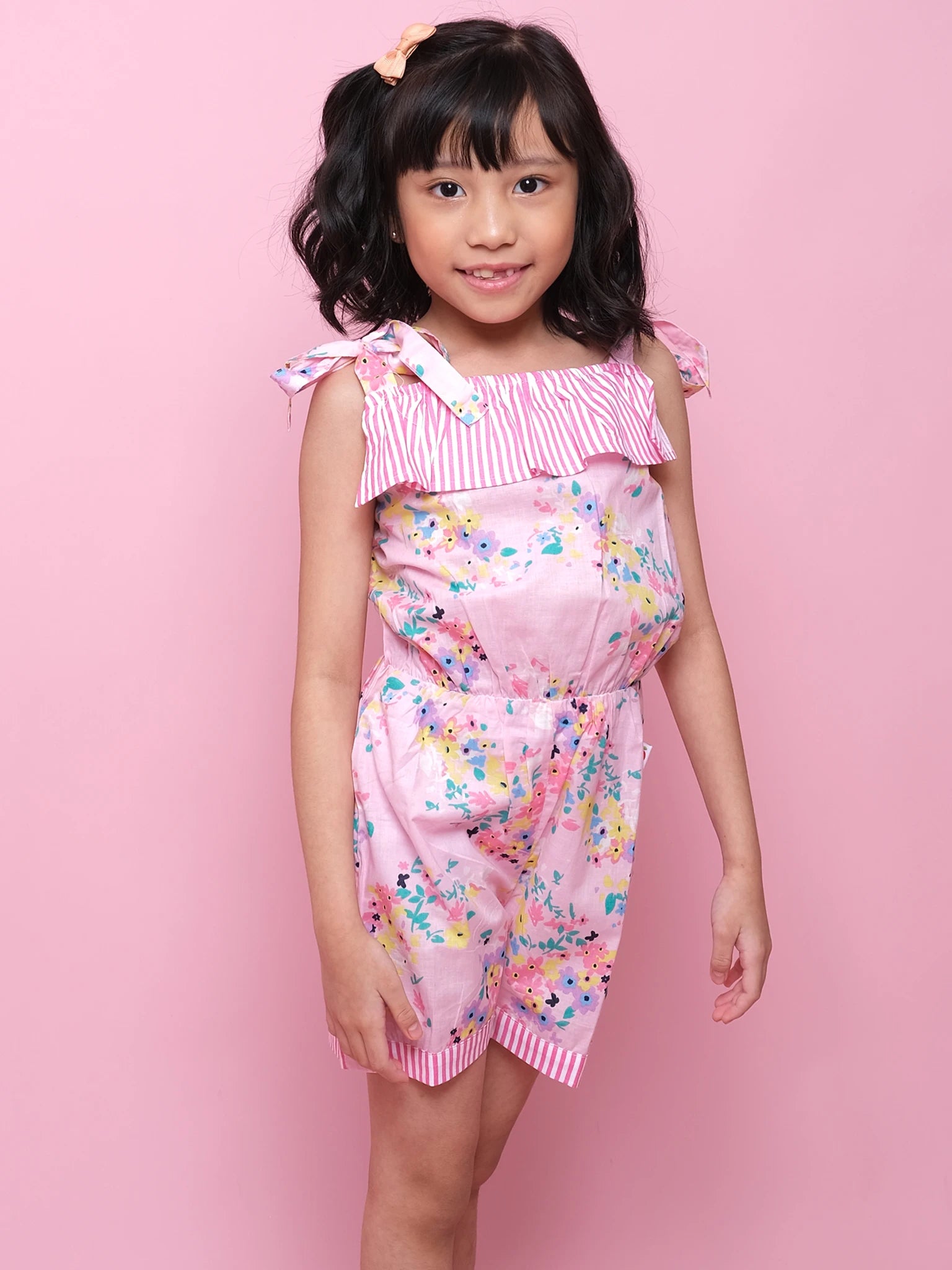 A young girl stands against a pink background wearing the Confetti Floral Josie Playsuit by Confetti Floral, featuring a pink floral cotton design with striped ruffle details and shoulder ties—perfect for summer.