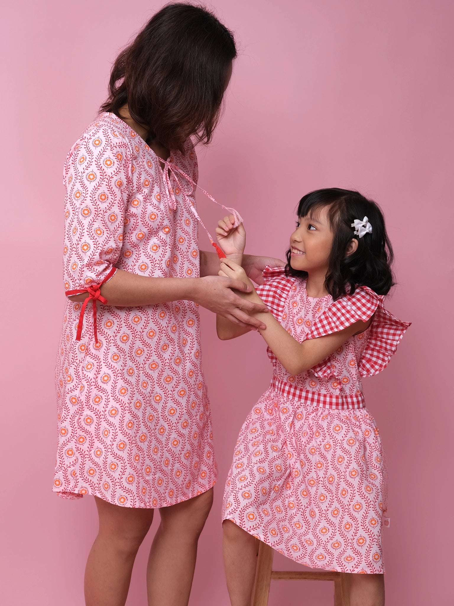 A woman and a girl wear matching Woodblock Women’s Resort Dresses by Woodblock Flower, featuring pink patterns inspired by woodblock prints, standing together against a pink background as the girl holds a beaded necklace.