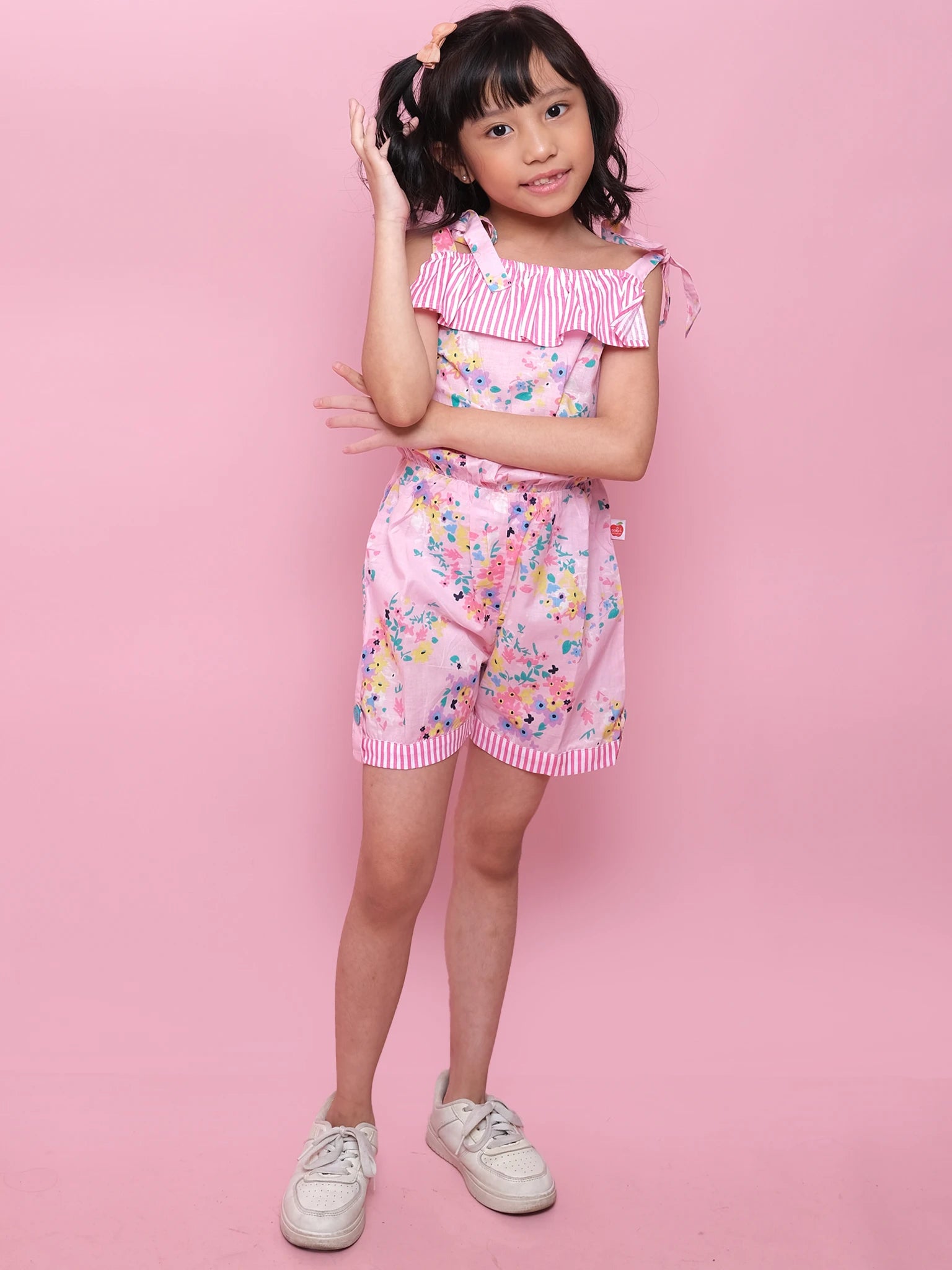 A young girl poses against a pink background in the Confetti Floral Josie Playsuit by Confetti Floral—a cotton floral playsuit styled with white sneakers, hand in hair and arm across her waist for an ideal summer look.