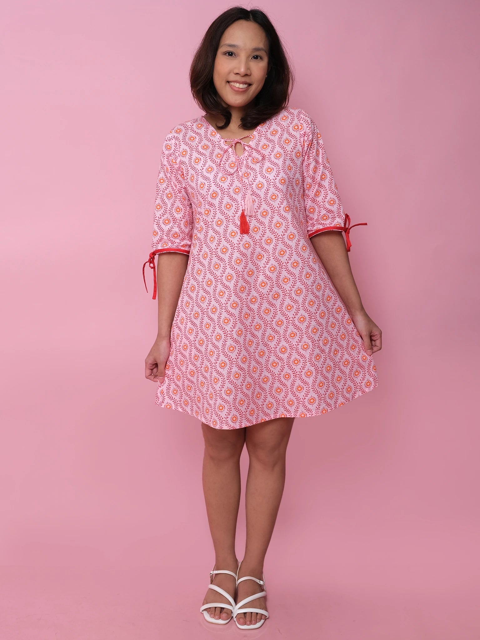 A woman stands against a pink background wearing the Woodblock Women’s Resort Dress by Woodblock Flower, featuring red tie sleeves and white sandals—a stylish update for any women’s wardrobe.
