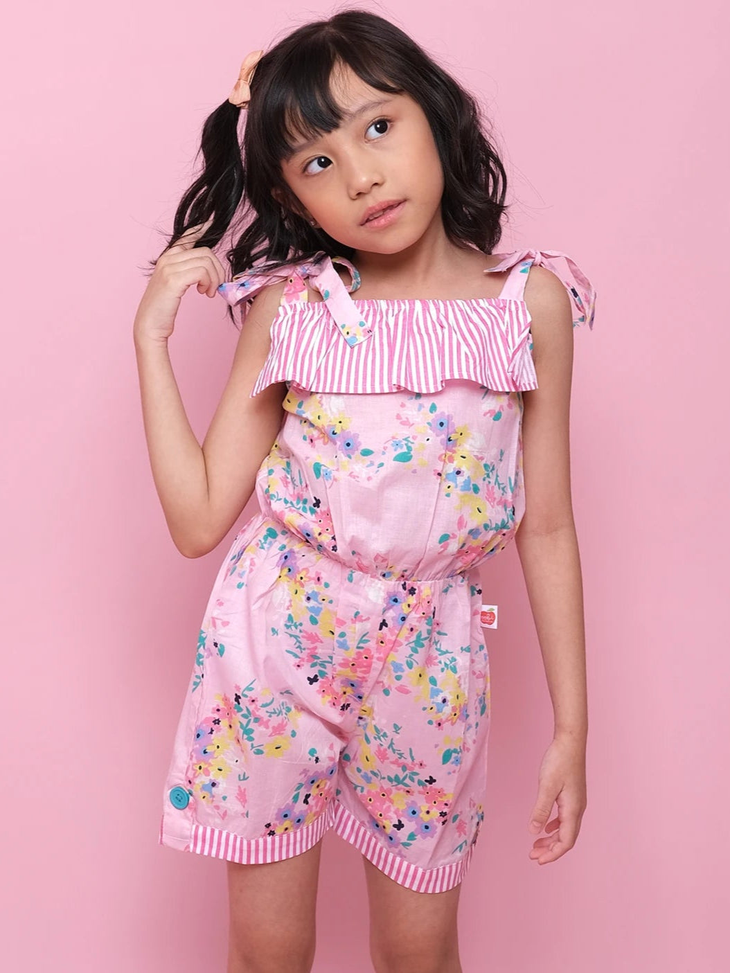 A young girl stands against a pink background in the Confetti Floral Josie Playsuit by Confetti Floral, featuring pink floral cotton fabric with striped details, paired with white sneakers and touching her hair.