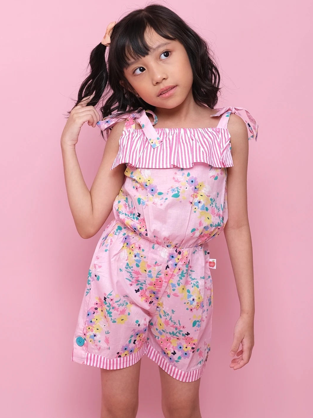 A young girl stands against a pink background, wearing the Confetti Floral Josie Playsuit with striped ruffle details and white sneakers, looking slightly to the side with one hand in her hair.