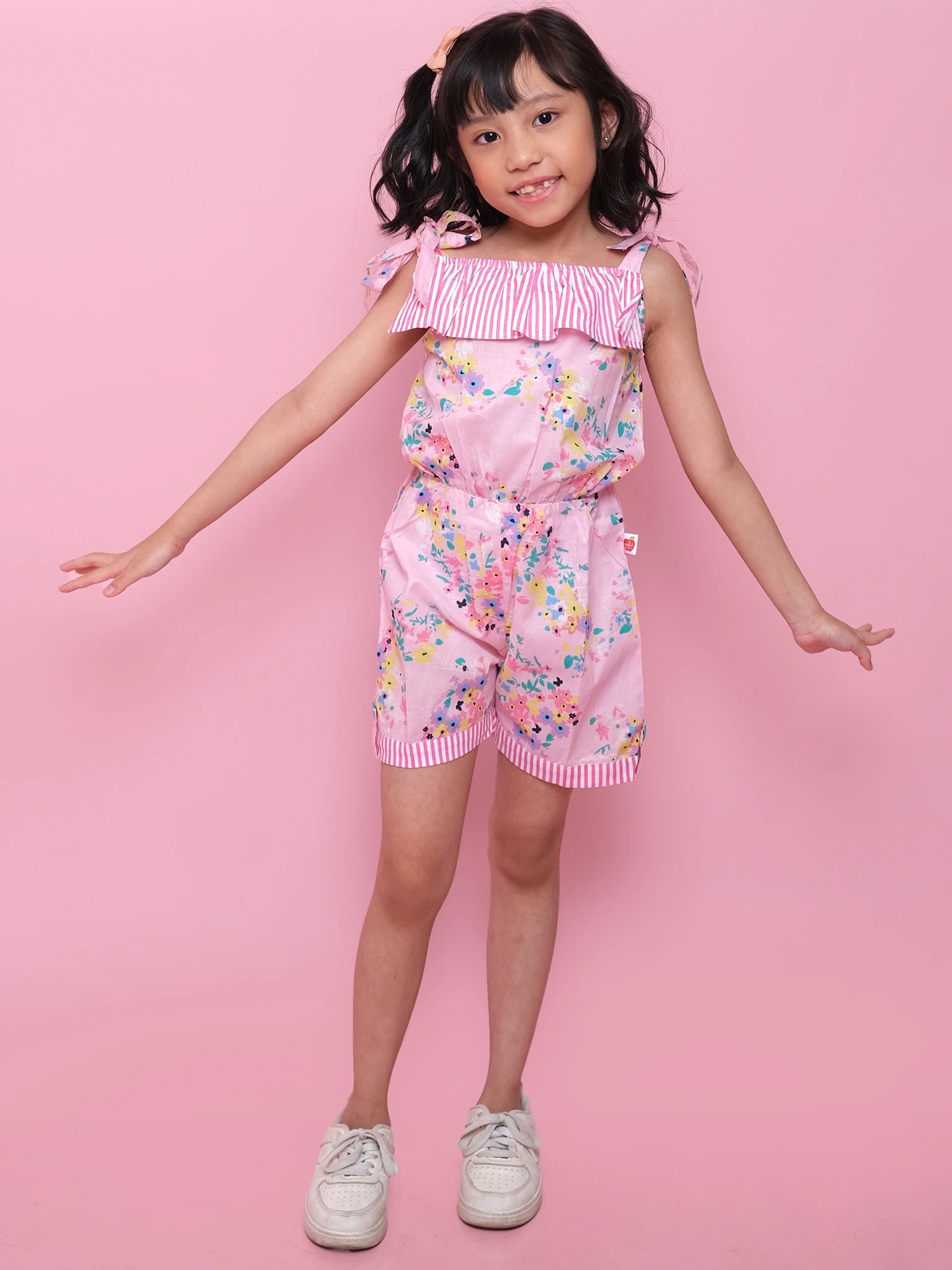 A young girl poses against a pink background, wearing a Confetti Floral Josie Playsuit made from lightweight cotton with striped ruffle details and white sneakers.