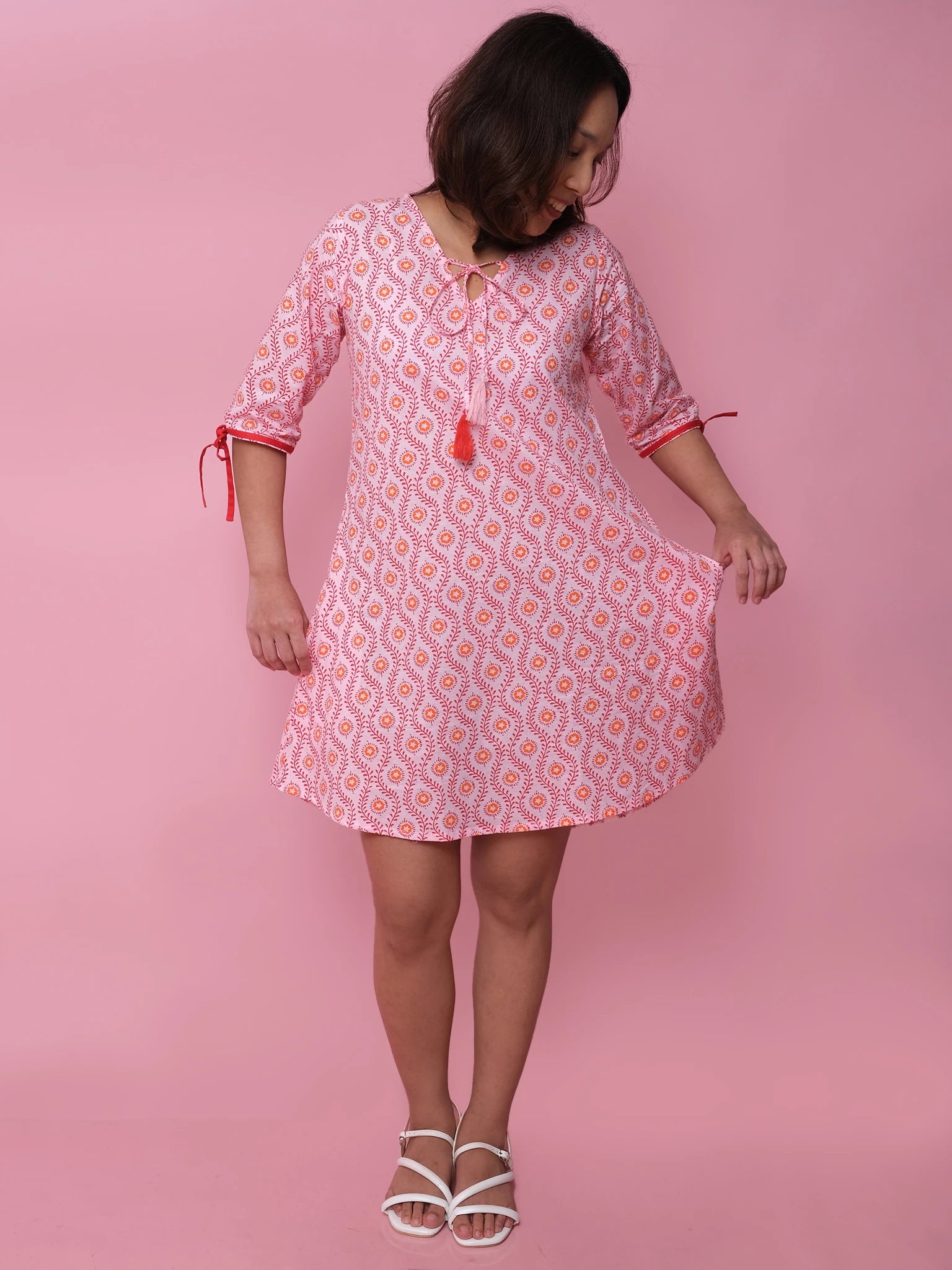 A woman wearing the Woodblock Women's Resort Dress by Woodblock Flower stands before a pink background, looking down as she holds part of her pink and white patterned kaftan dress with red tassels, paired with white sandals.