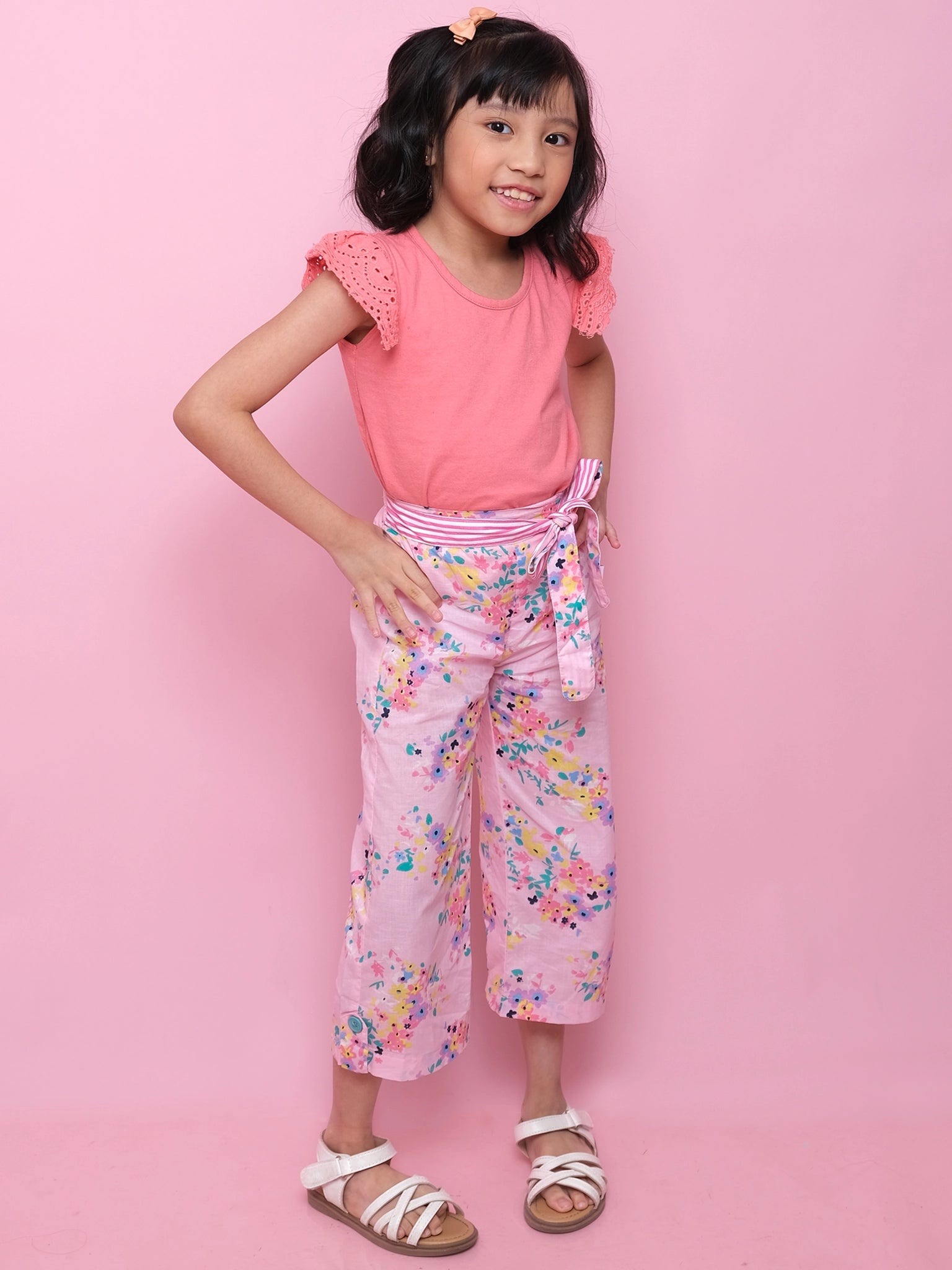 A young girl stands against a pink background in Confetti Floral Wide-Leg Pants, featuring an elastic waist, striped sash, and floral print, paired with a coral top and white sandals.