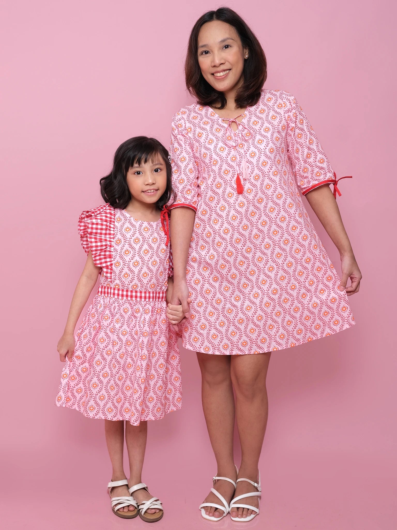An adult and a child hold hands against a pink backdrop, both in patterned Woodblock Flower Women's Resort Dresses paired with white sandals.