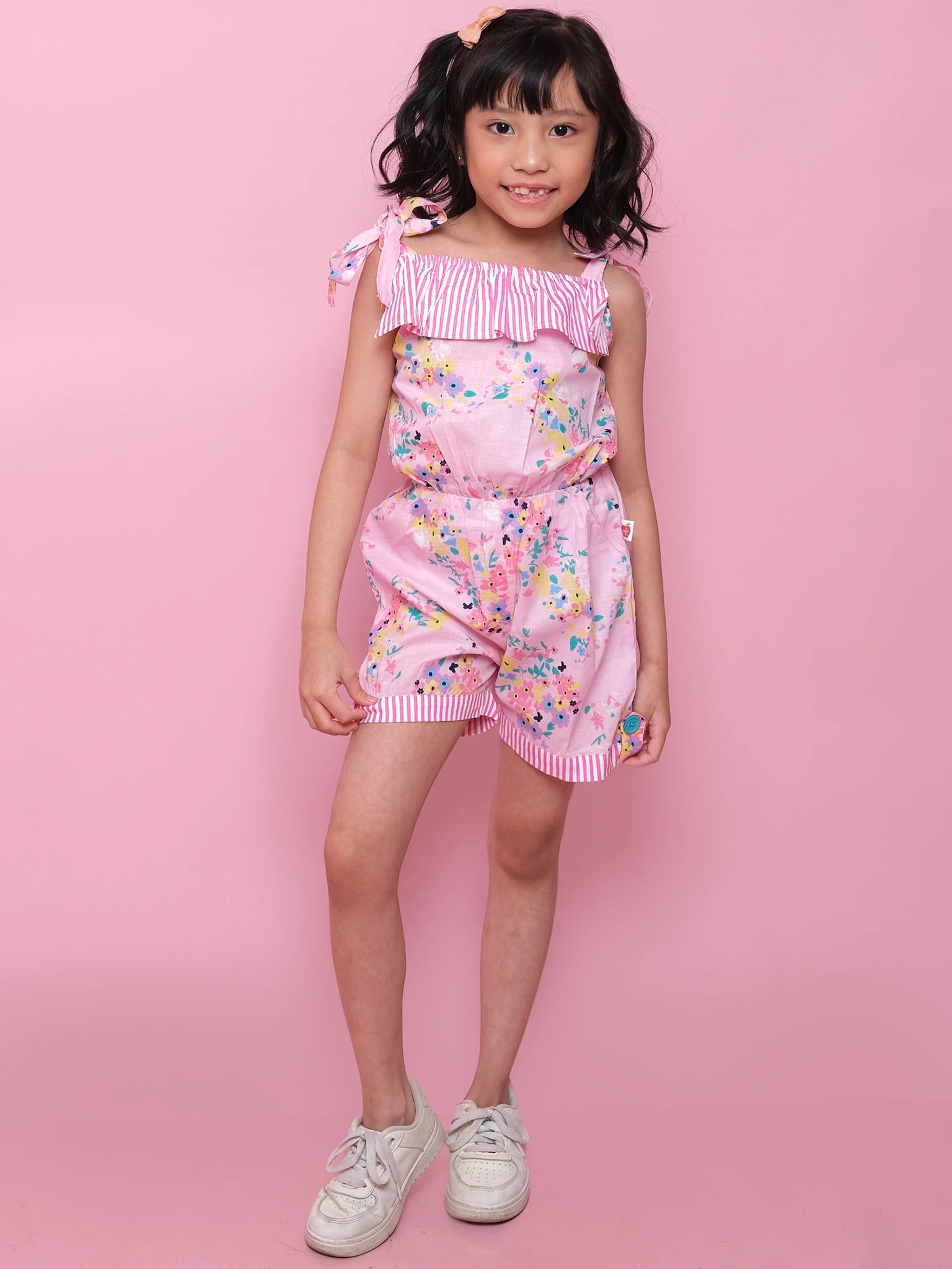 A young girl smiles at the camera in a Confetti Floral Josie Playsuit by Confetti Floral, featuring pink florals with striped details and crafted from lightweight cotton, styled against a pink background with white sneakers.