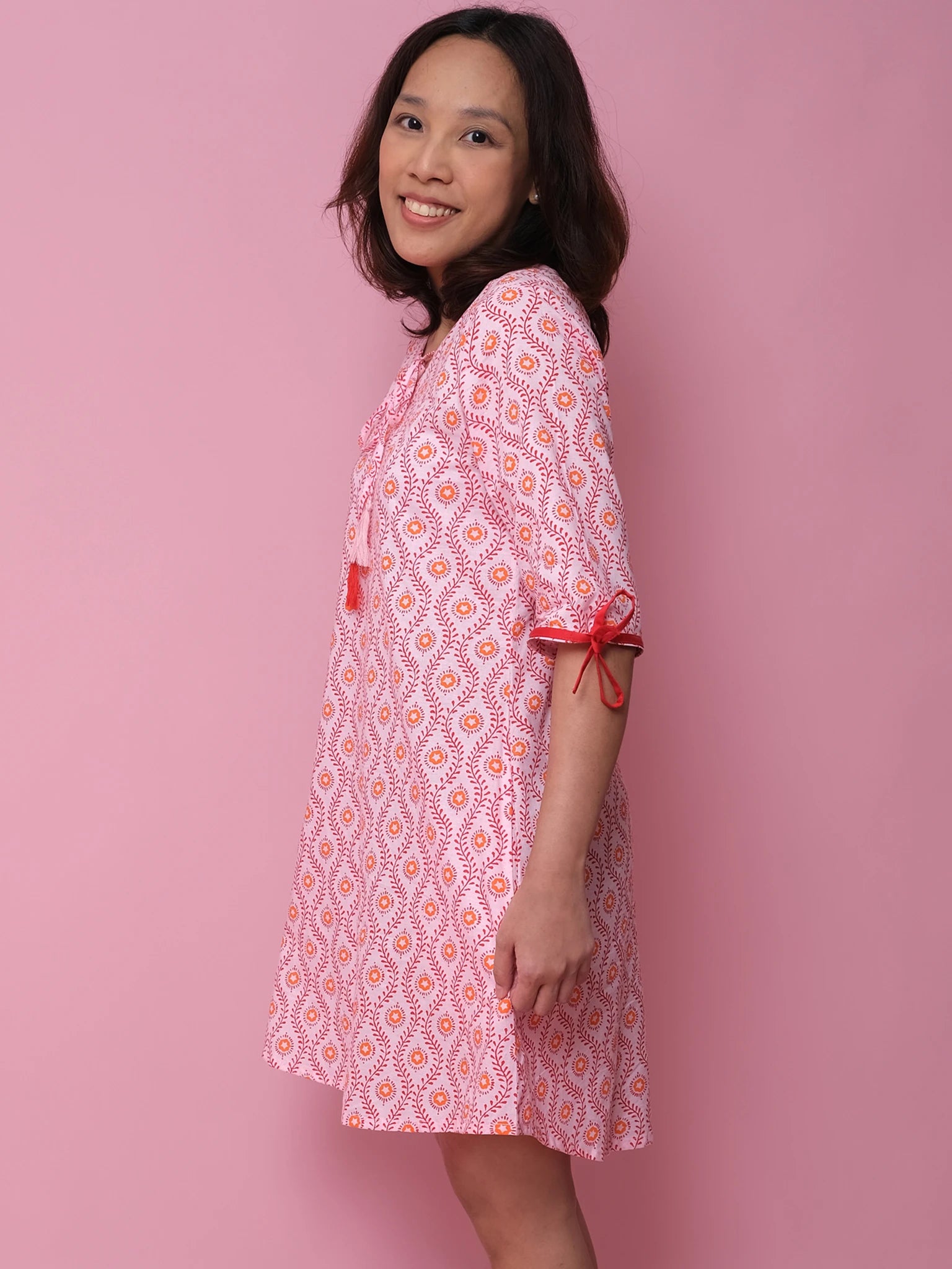 A woman smiles sideways against a pink background, wearing the Woodblock Flower Woodblock Women's Resort Dress—a loose, patterned kaftan with sleeve tie details and a charming woodblock print, perfect for stylish women's resort wear.