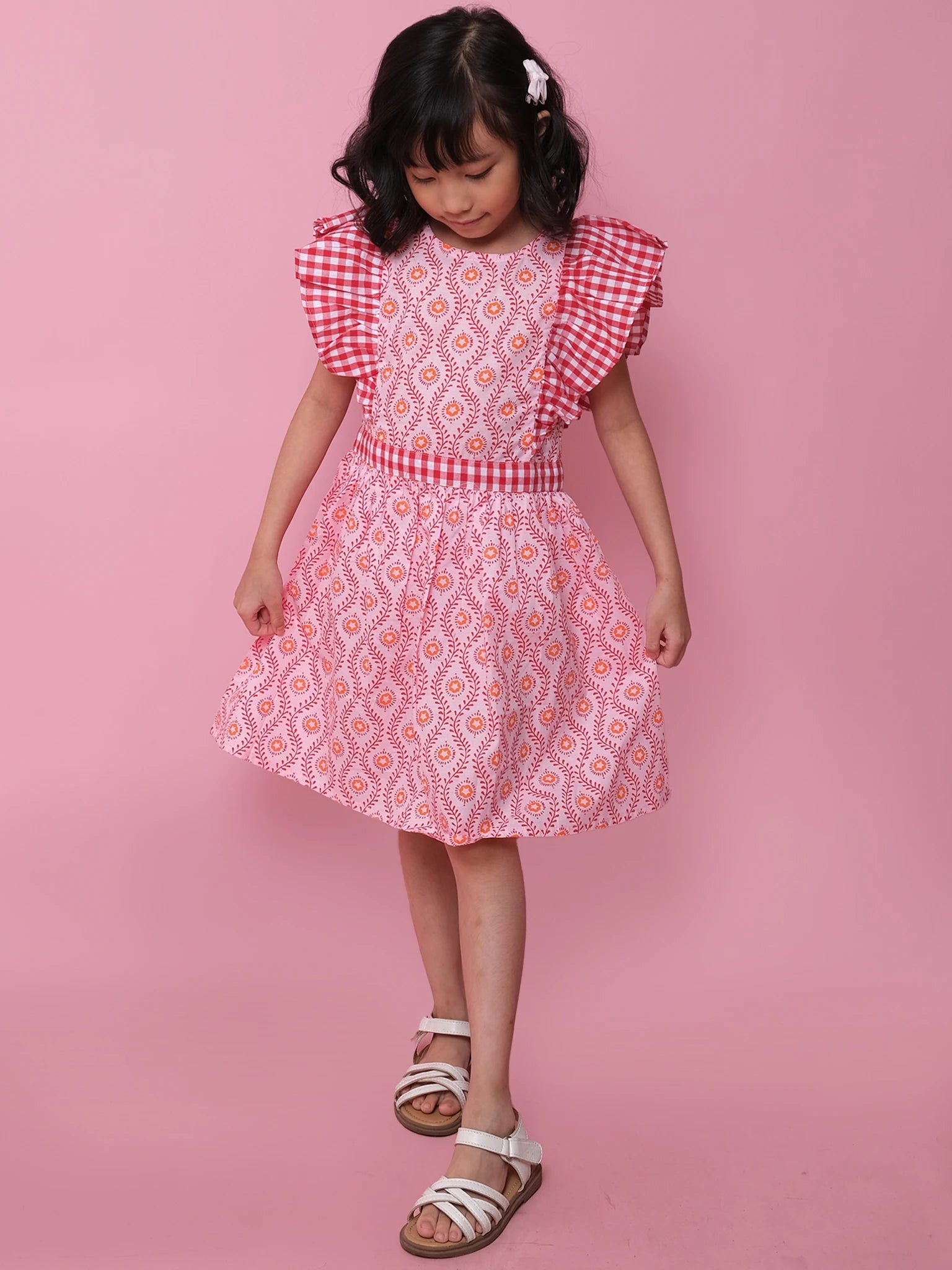 A young girl wears the Woodblock Flower Jayne Dress with ruffled gingham sleeves and white sandals, standing against a plain pink background.