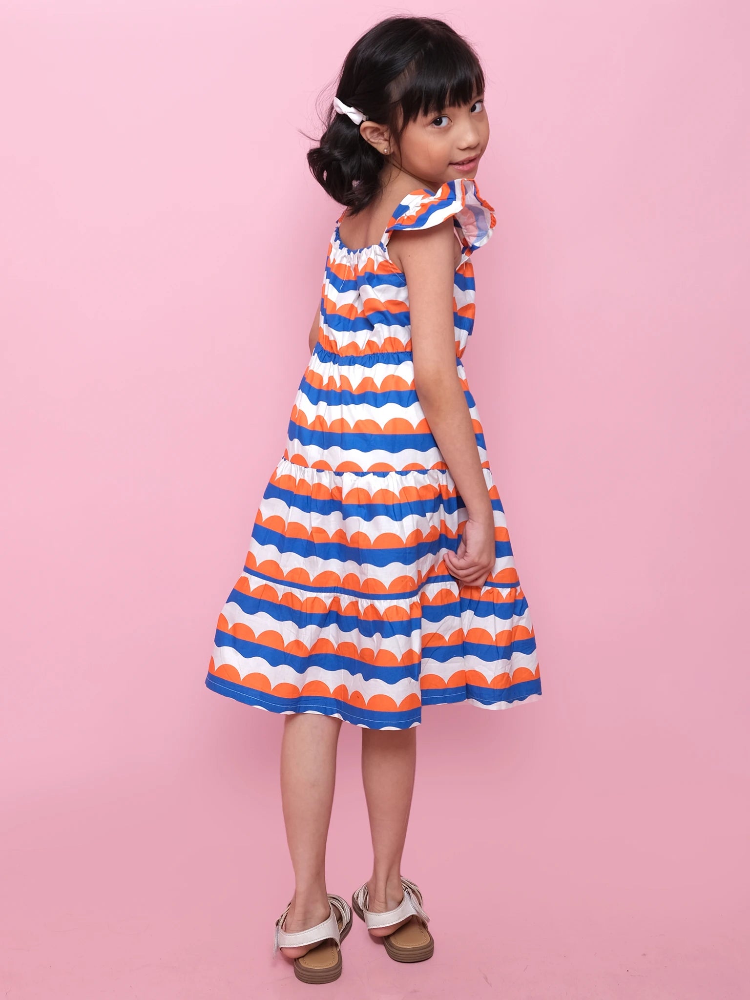 A young girl with dark hair in a clip wears the Nautical Holiday Dress by Nautical and sandals, standing against a pink background and looking over her shoulder.