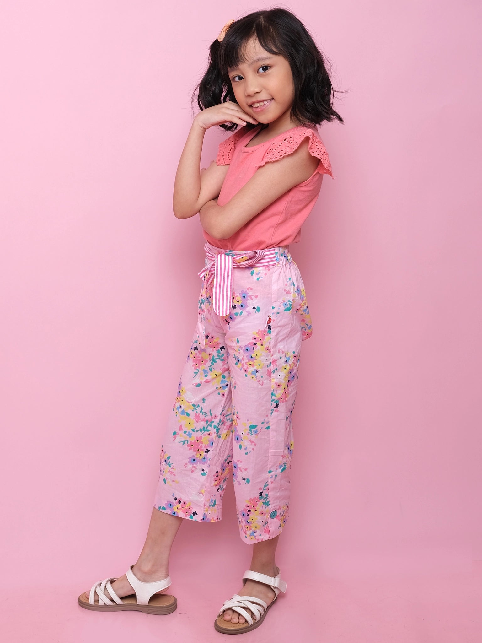 A young girl smiles with one hand on her cheek, wearing a pink top, Confetti Floral Wide-Leg Pants by Confetti Floral with an elastic waist, and white sandals against a pink background.