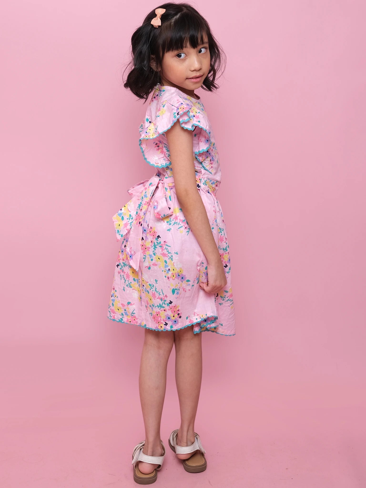 A young girl with short dark hair stands sideways against a pink background, wearing the Confetti Floral Lacy Dress by Confetti Floral, paired with a hair clip and white sandals.