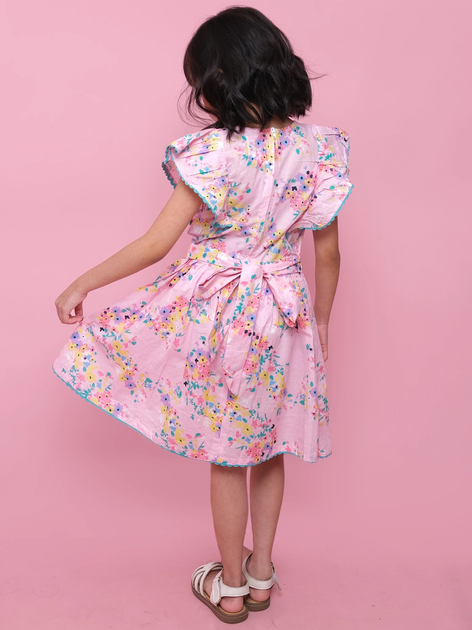 A girl poses against a pink background in the Confetti Floral Lacy Dress by Confetti Floral, featuring a light pink floral print, bow at the back, and white sandals, holding out one side of her skirt.