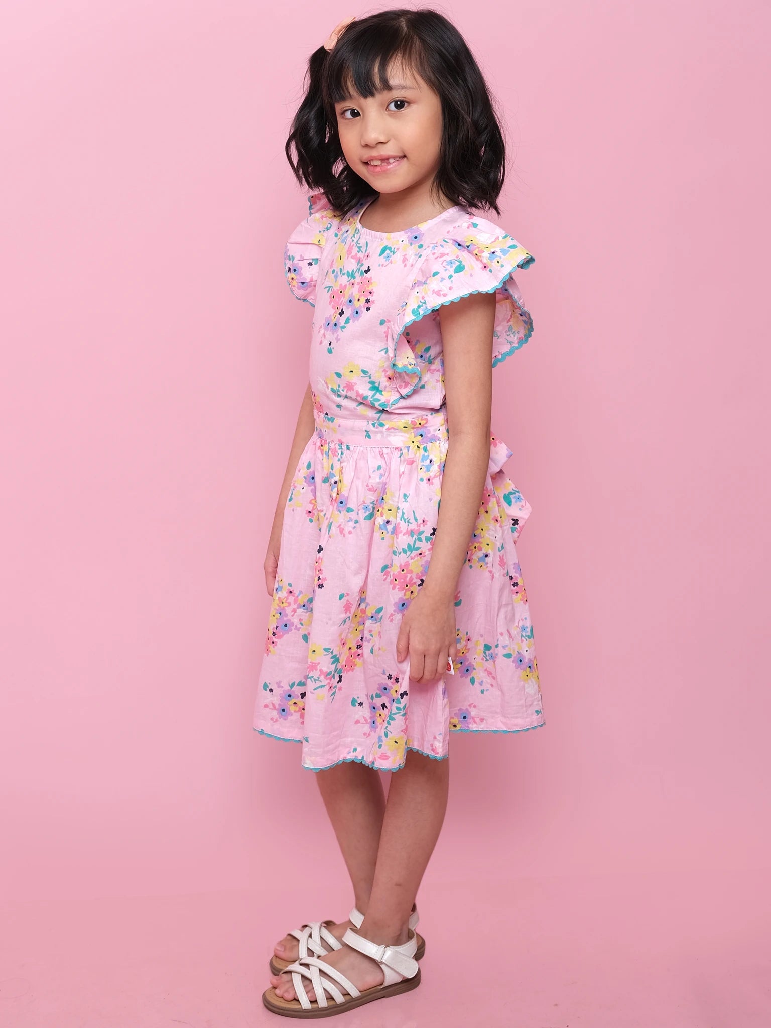 A young girl stands sideways against a pink background, wearing the Confetti Floral Lacy Dress by Confetti Floral, featuring pink floral lace, ruffled sleeves, and white sandals.