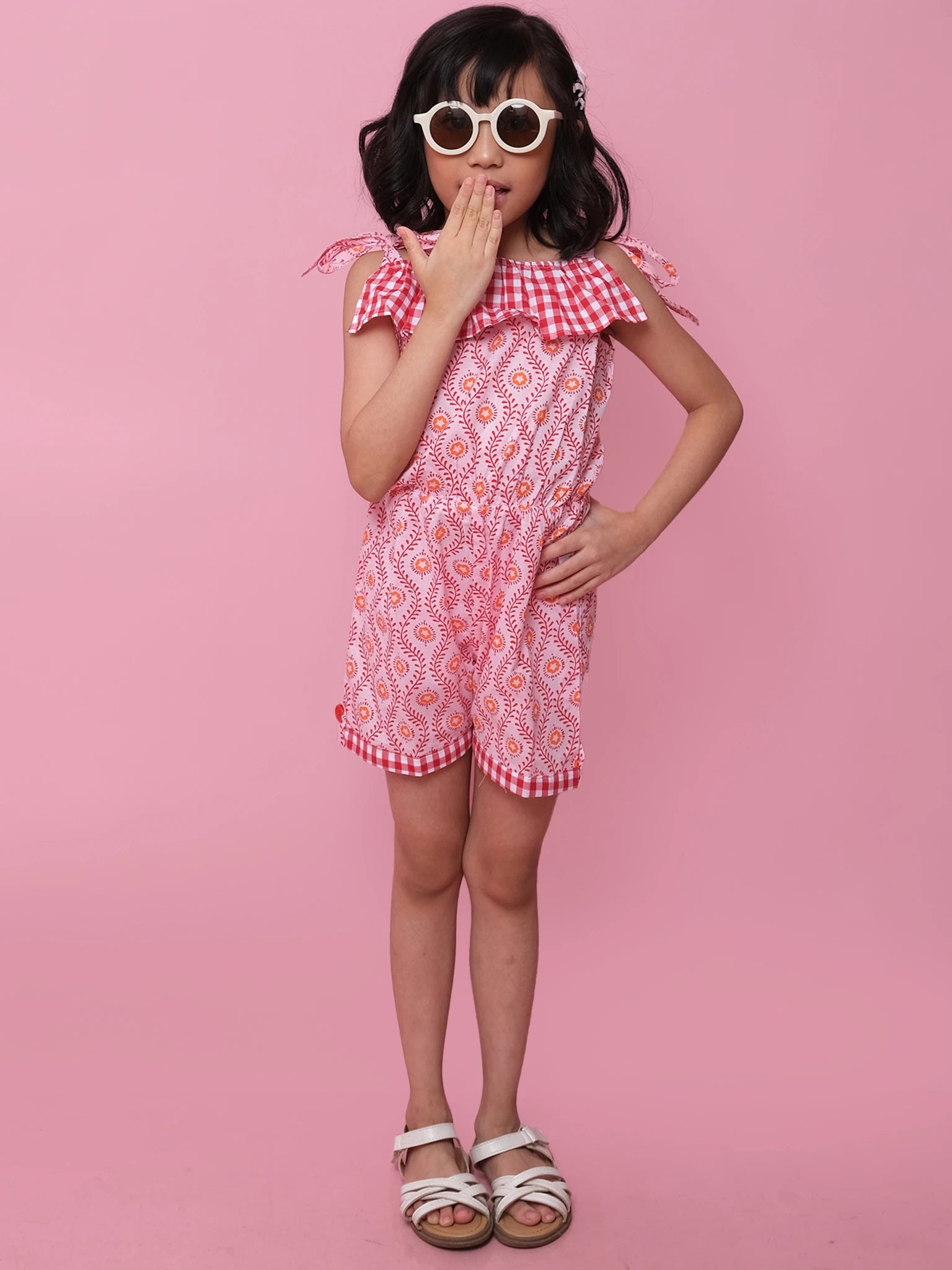 A young girl poses against a pink background wearing sunglasses, white sandals, and the Woodblock Flower Josie Playsuit by Woodblock Flower, her hand on her hip and the other covering her mouth.