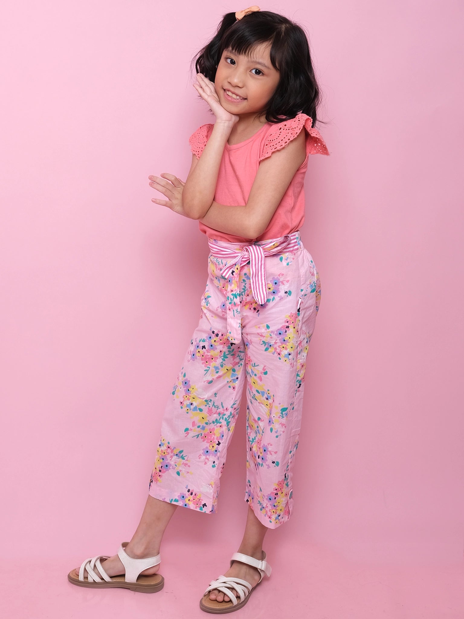 A young girl poses against a pink background wearing a pink top, Confetti Floral Wide-Leg Pants by Confetti Floral, and white sandals, with one hand on her cheek and the other on her arm.