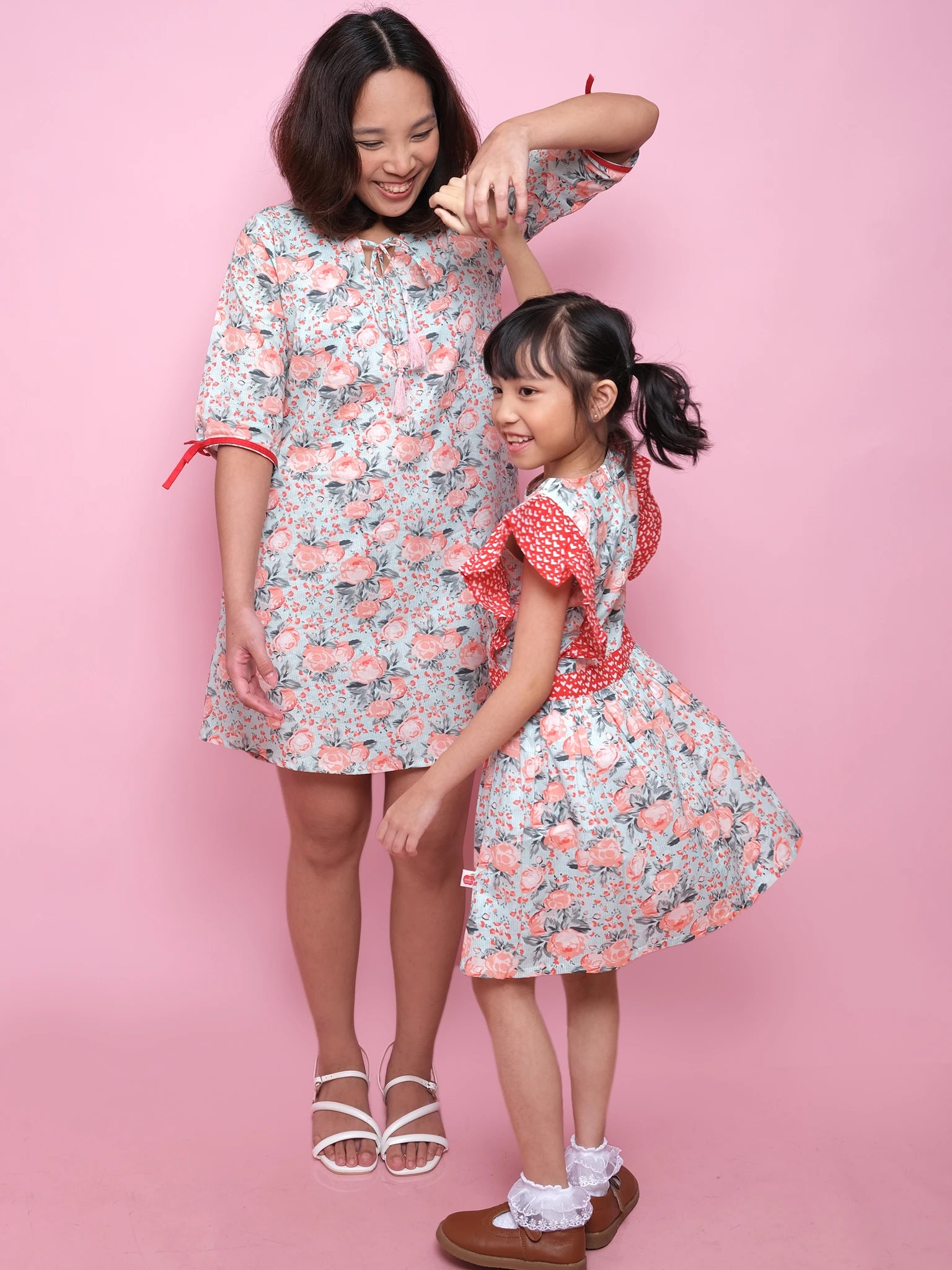 An adult and child, both in Vintage Roses Jayne Dresses, stand against a pink backdrop; the adult smiles and twirls the child.