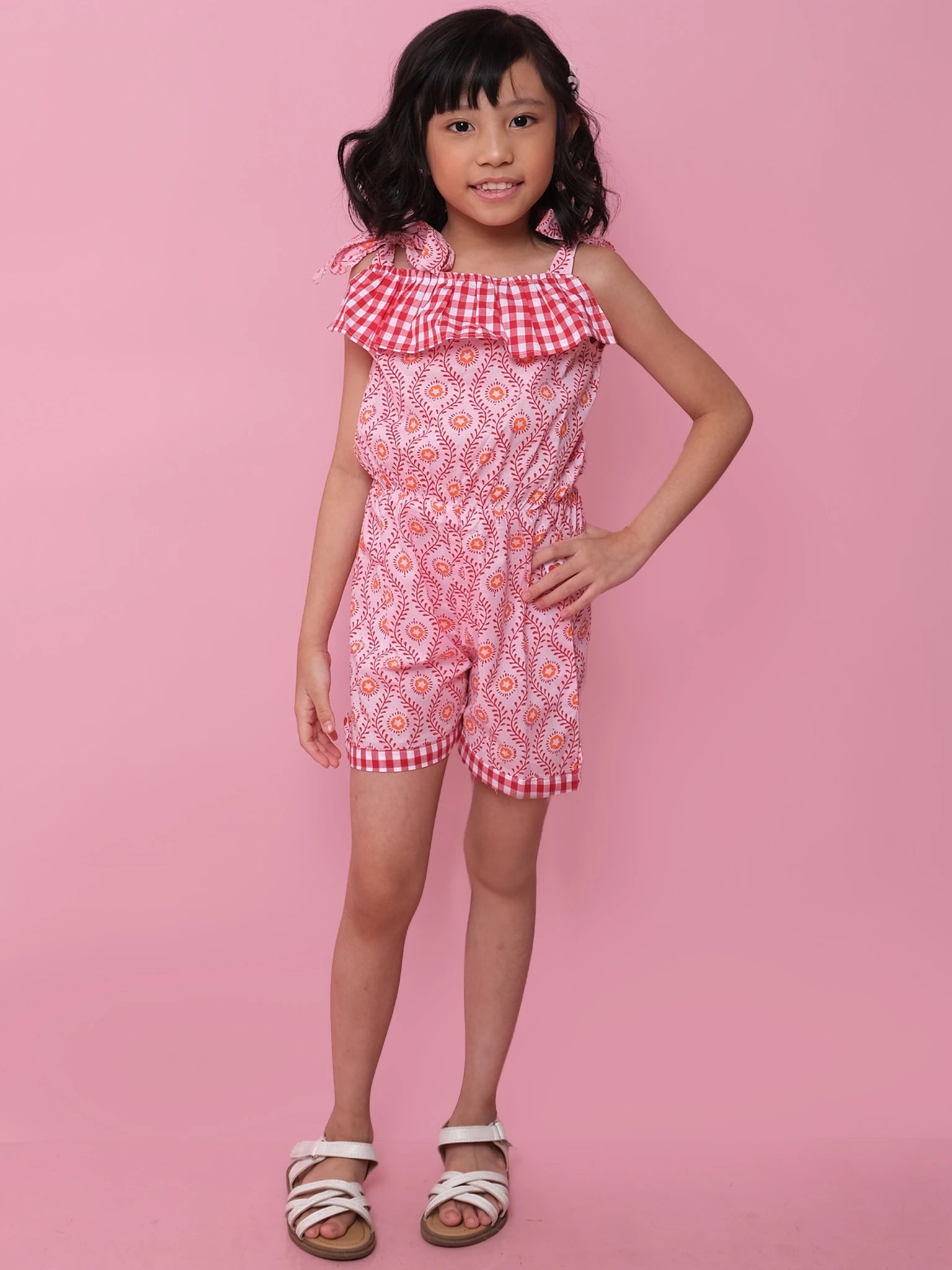 A young girl stands against a pink background wearing the ethically made Woodblock Flower Josie Playsuit in red and white with ruffle details, paired with white sandals.