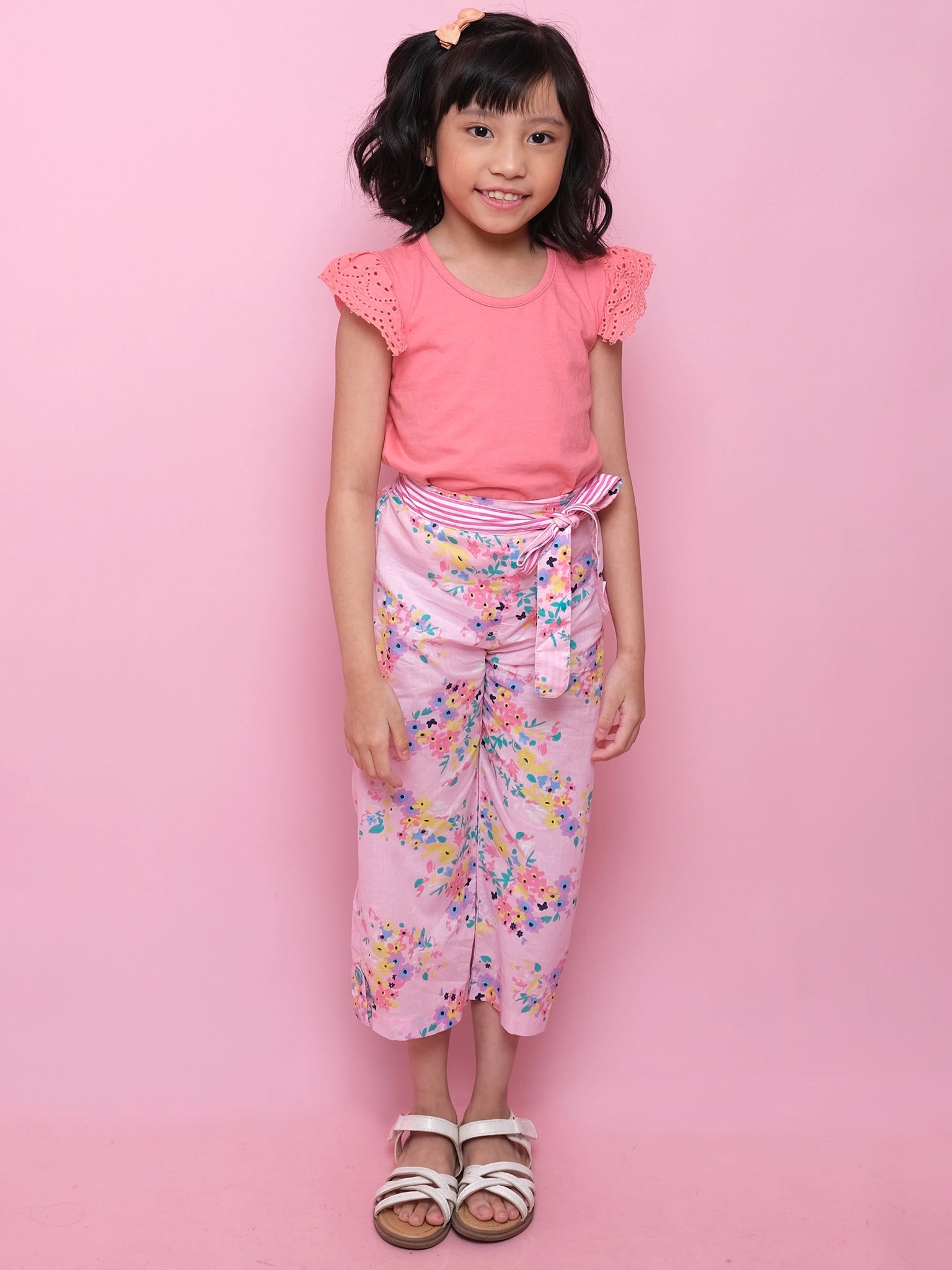 A young girl stands against a pink background, wearing Confetti Floral Wide-Leg Pants by Confetti Floral with an elastic waist and sash, paired with a coral lace-sleeve top and white sandals.