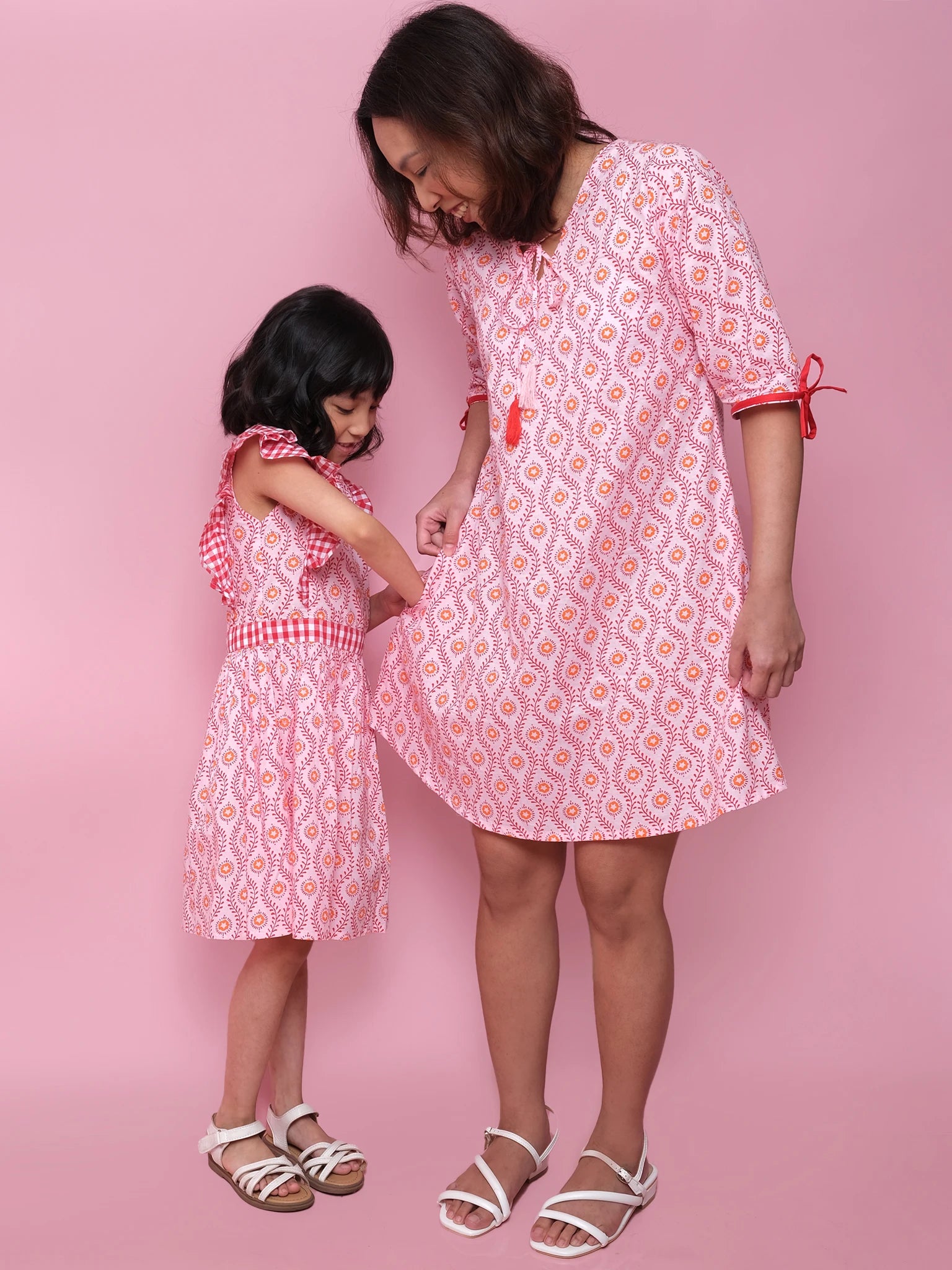 A woman and child wear matching patterned pink and white Woodblock Women's Resort Dresses by Woodblock Flower, paired with white sandals, standing against a pink background—a lovely display of coordinated style.