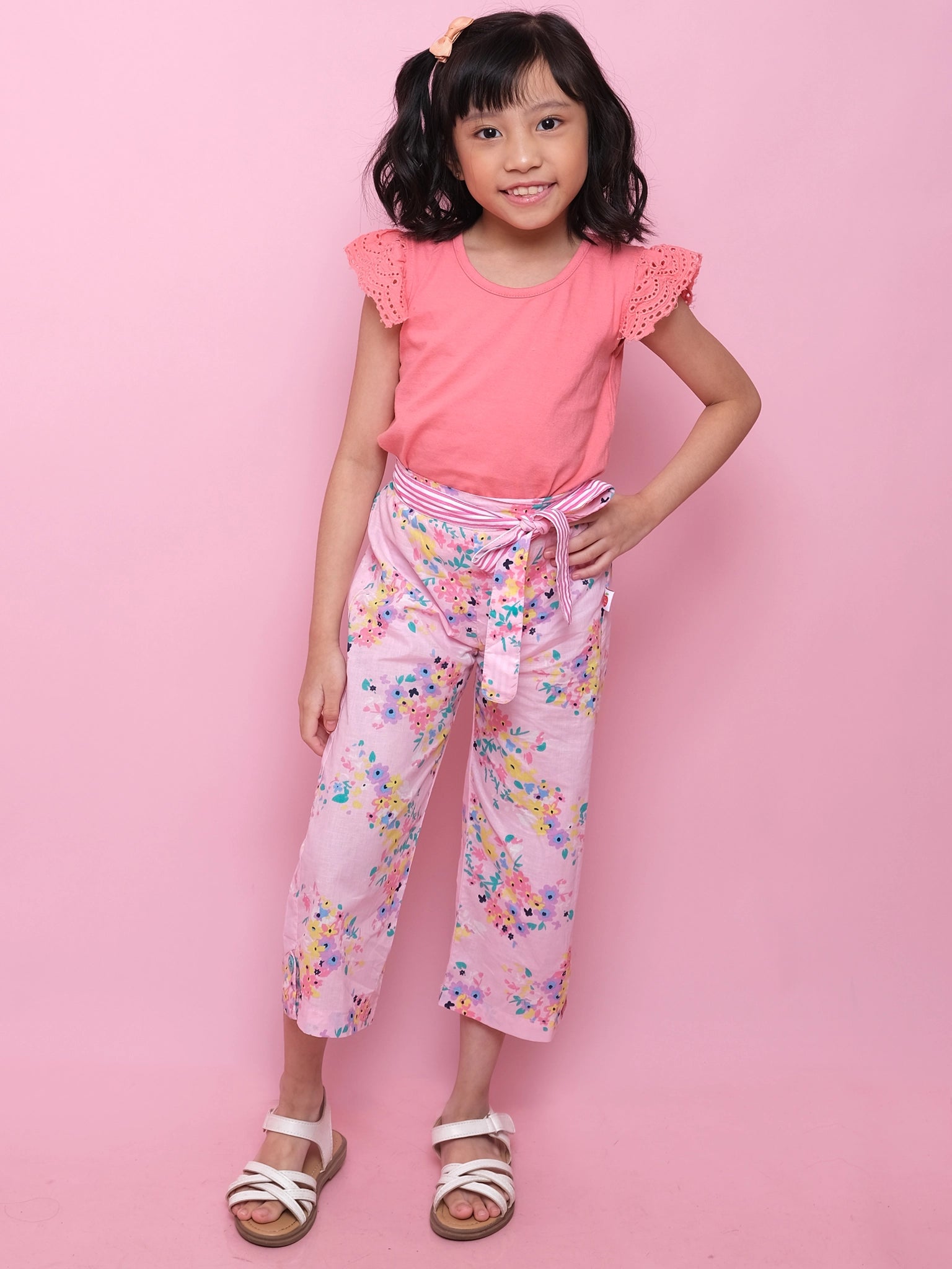 A young girl smiles with one hand on her hip, wearing Confetti Floral Wide-Leg Pants from Confetti Floral, paired with a pink top, white sandals, and a hair clip against a pink background.
