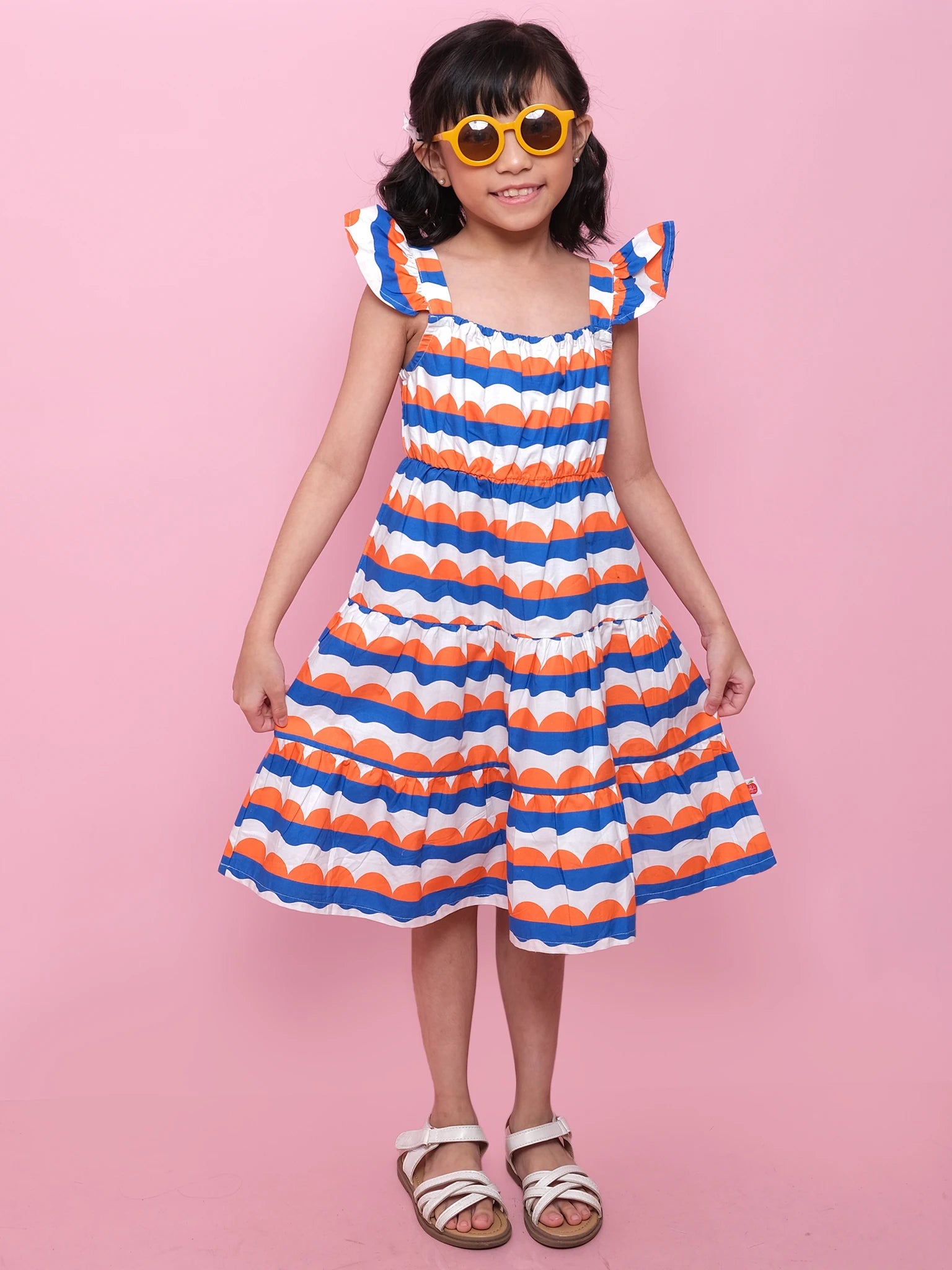 A young girl stands against a pink background wearing the Nautical Holiday Dress by Nautical—a ruffled, sleeveless seaside dress with blue and orange scallop stripes—paired with white sandals and yellow round sunglasses.