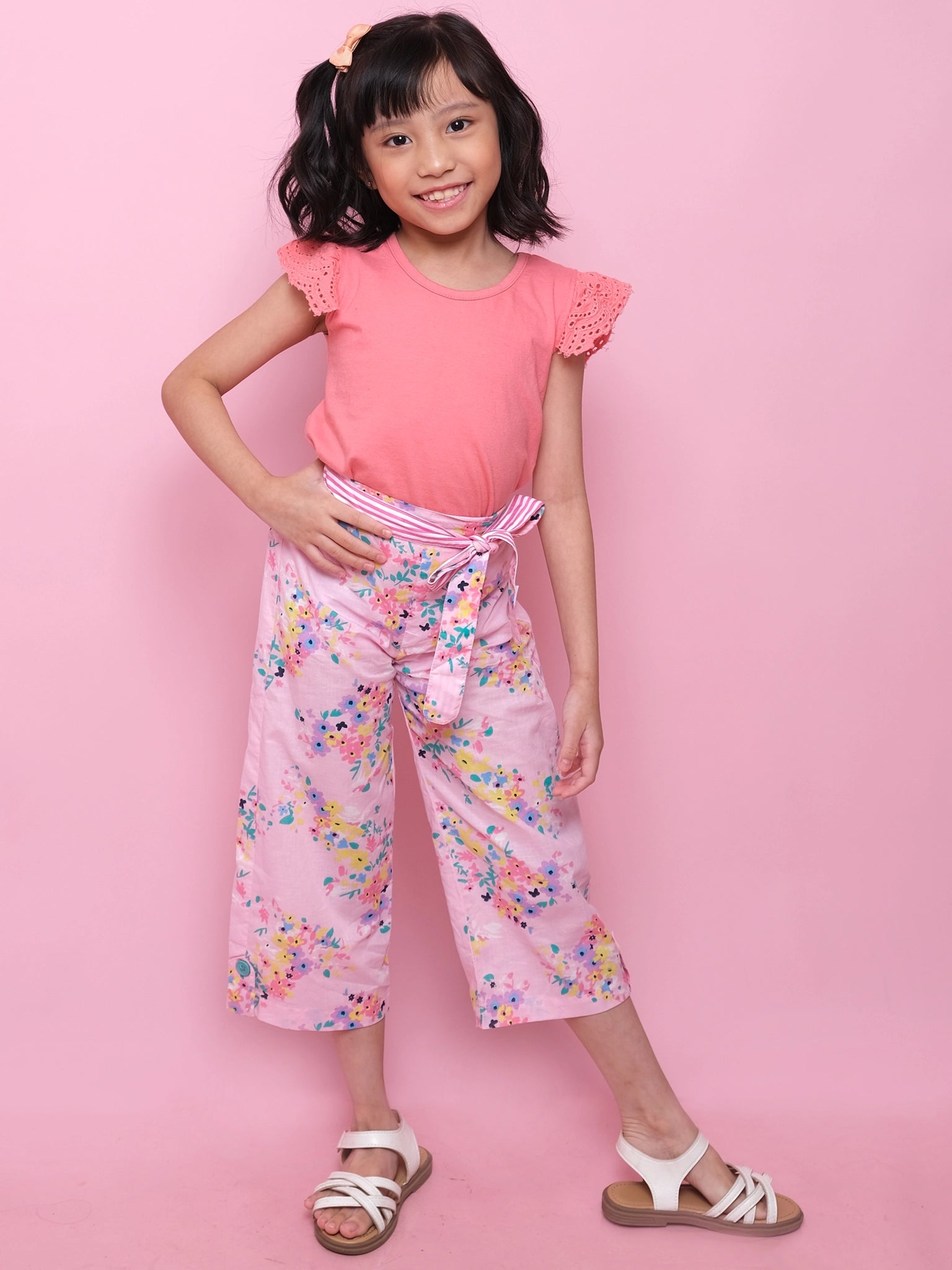 A young girl smiles at the camera against a pink background, wearing Confetti Floral Wide-Leg Pants, a pink top, white sandals, and a hair clip.