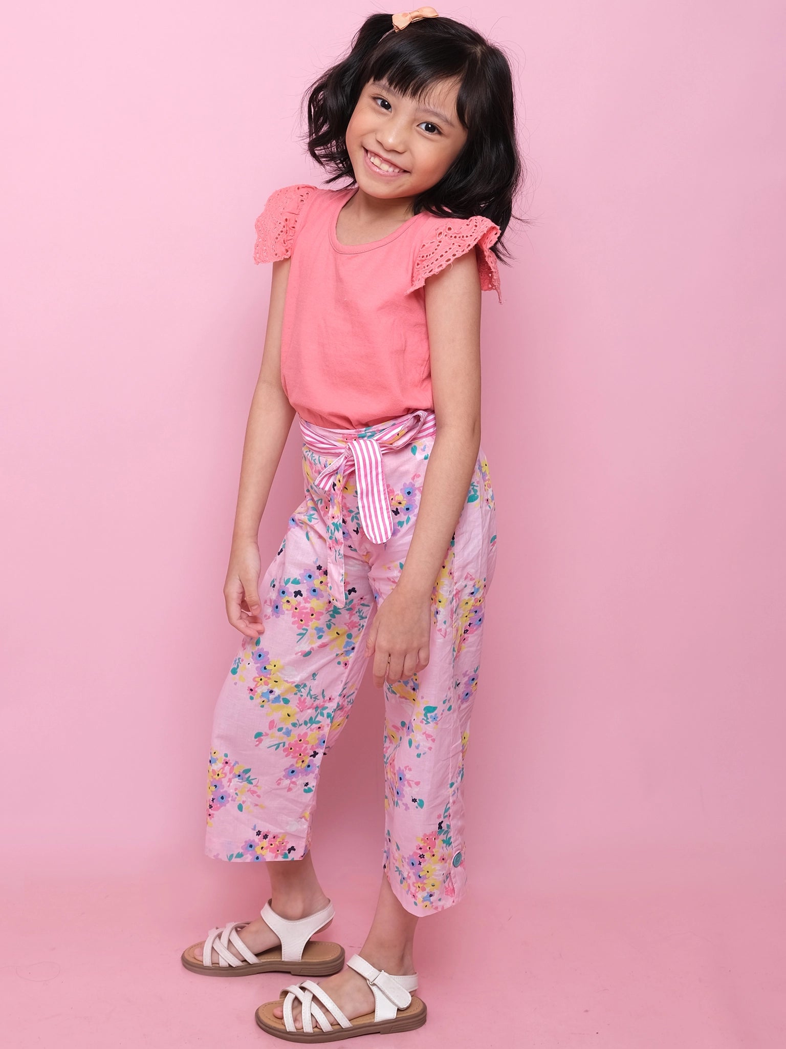 A young girl stands against a pink background, wearing Confetti Floral Wide-Leg Pants with hand-printed pink florals, an elastic waist, striped sash, and a peach top, paired with white sandals.