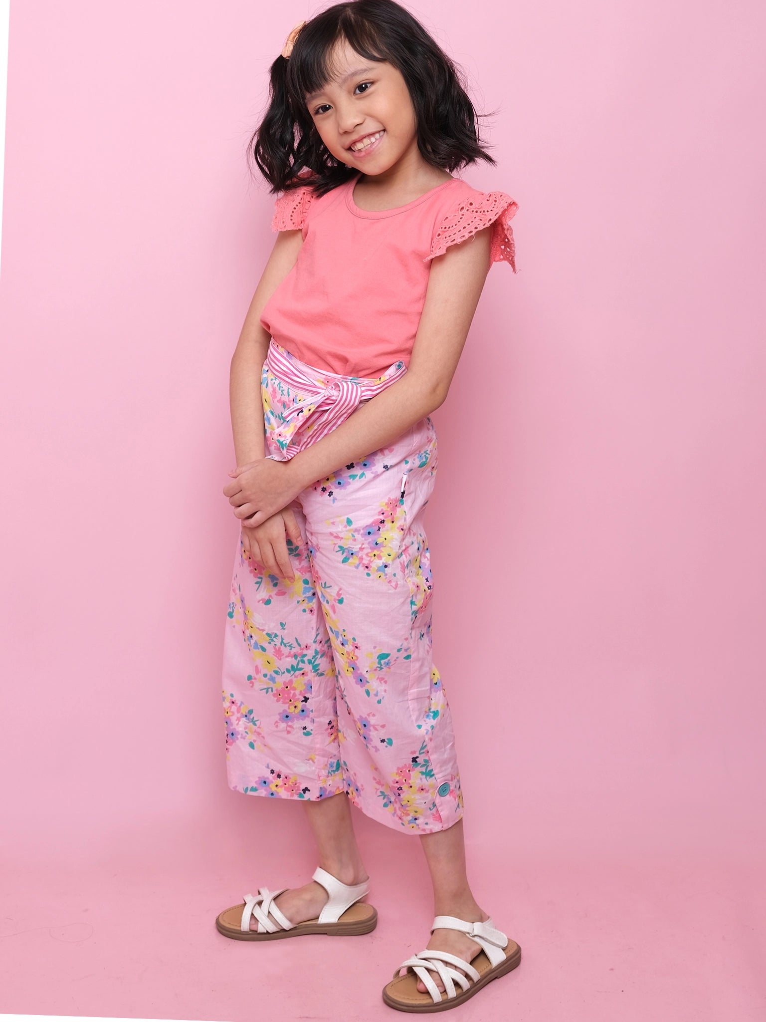 A young girl smiles against a pink backdrop, wearing Confetti Floral Wide-Leg Pants by Confetti Floral with an elastic waist, paired with a pink top and white sandals.