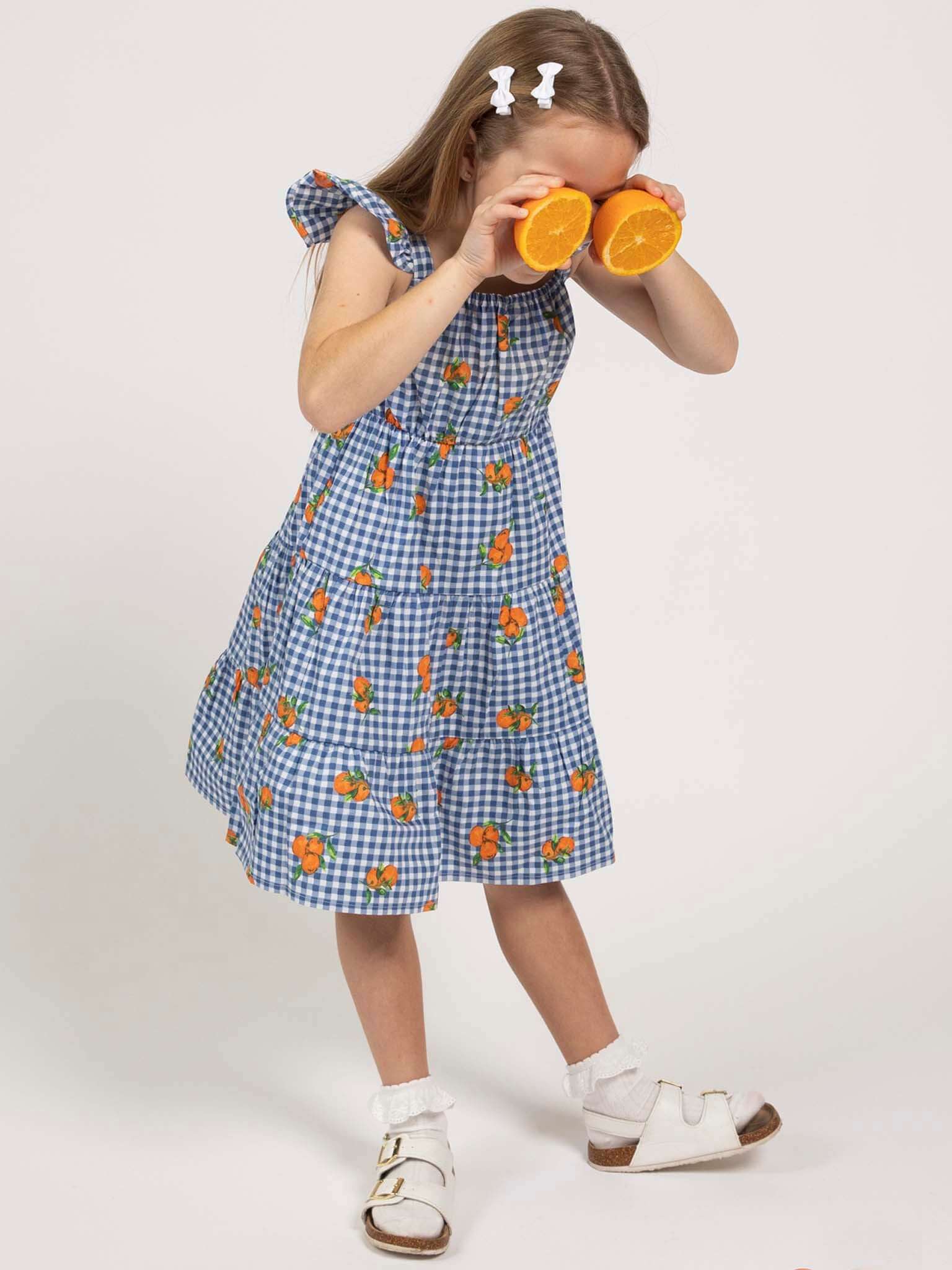 A young girl in the Amalfi Oranges Holiday Dress by Amalfi Oranges, a blue gingham summer dress with orange patterns, playfully holds two orange halves over her eyes against a plain background.