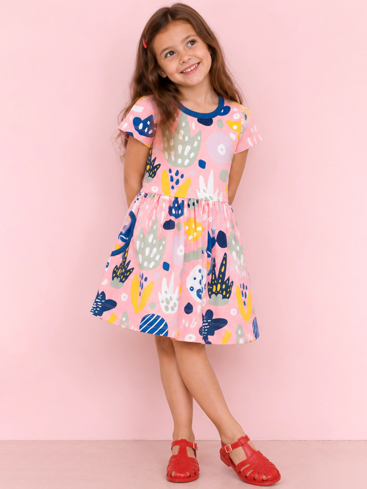 A young girl with long brown hair stands against a solid pink background wearing a short-sleeved pink dress with a colorful abstract pattern and red sandals.