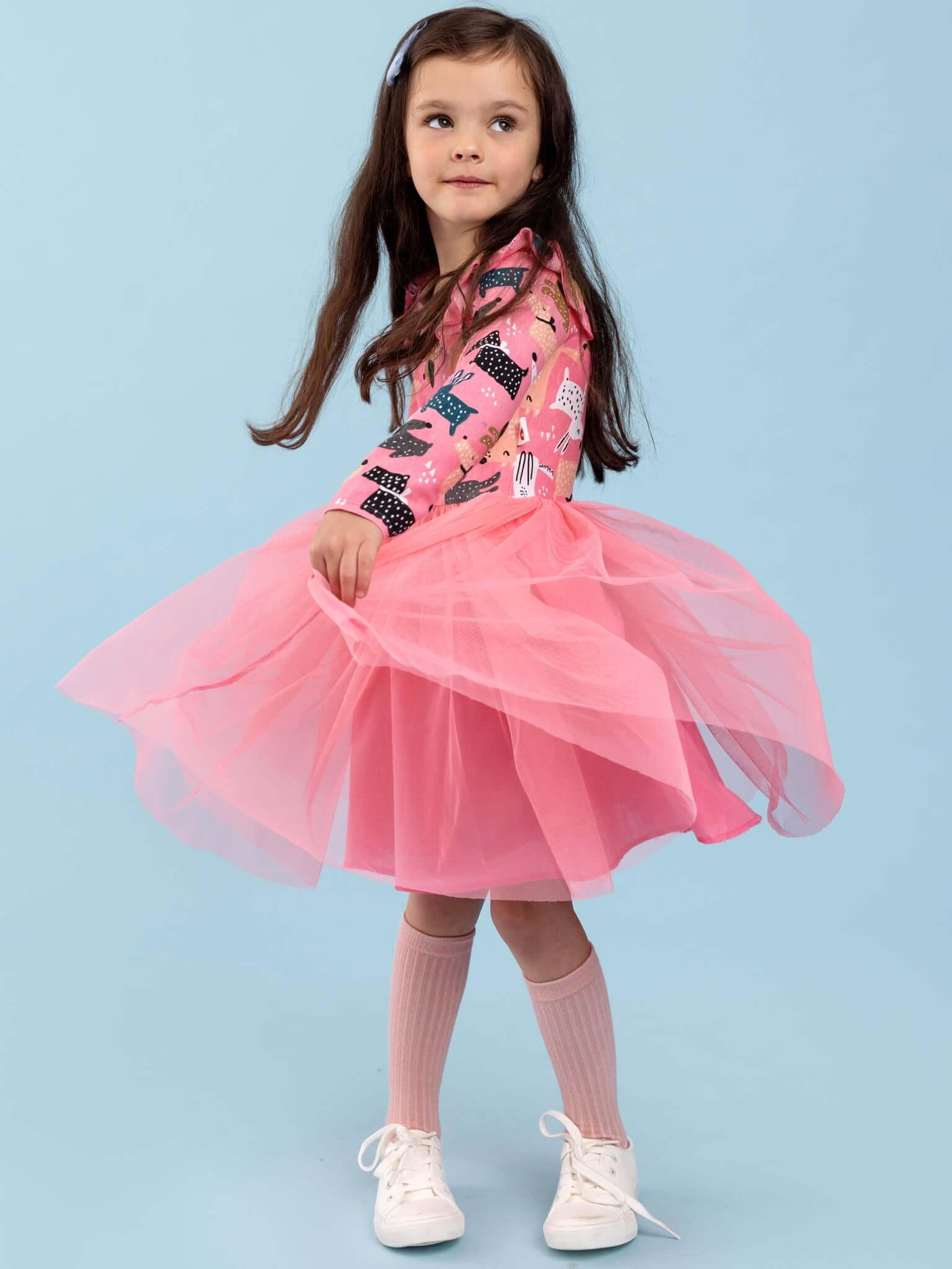 A young girl models the Oobi Ariel Test outfit—pink tulle skirt and pink patterned long-sleeve top—paired with knee-high socks and white shoes, standing before a plain light blue background.