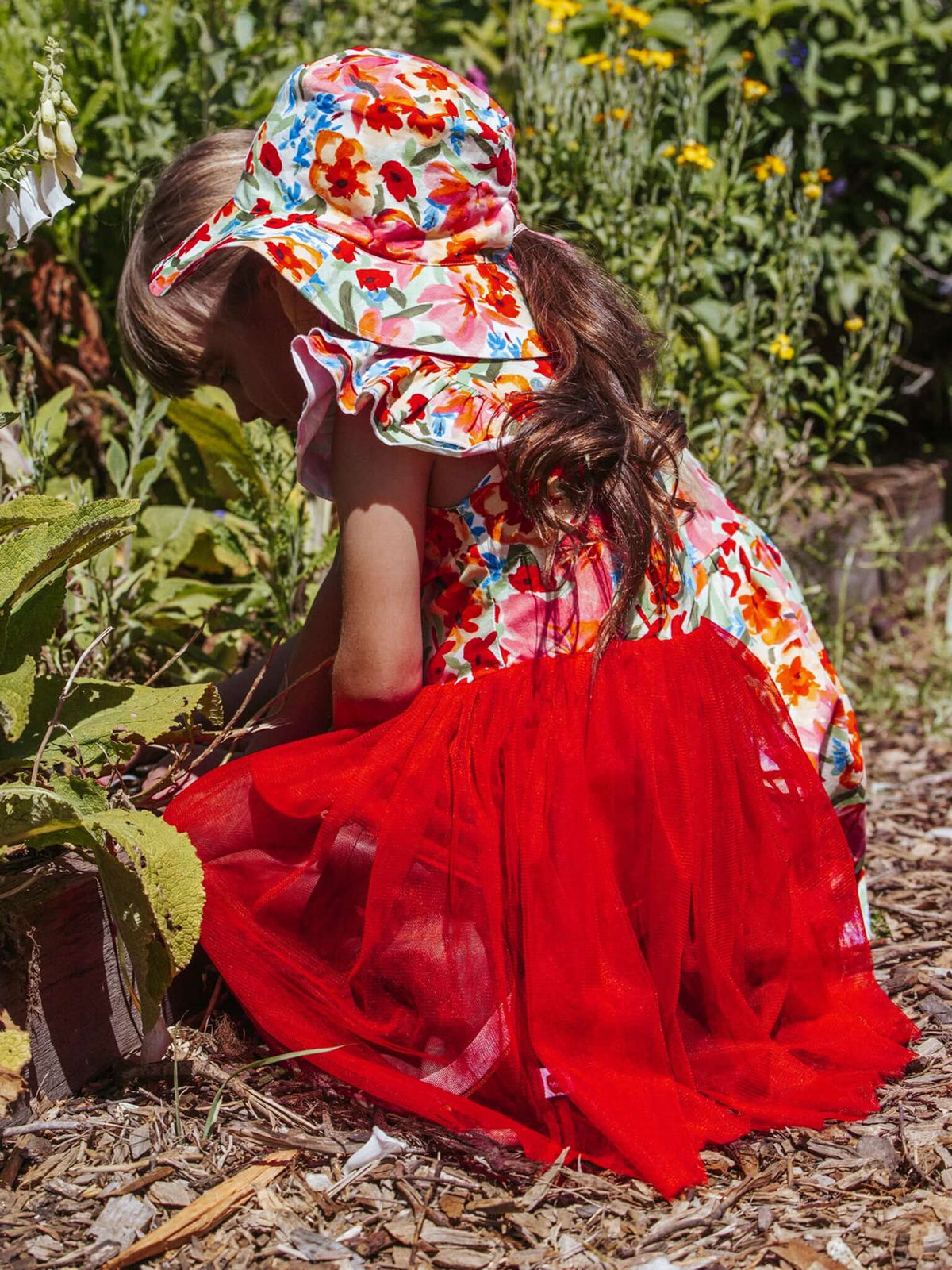 Child in a colorful floral hat and red dress sitting in a garden.