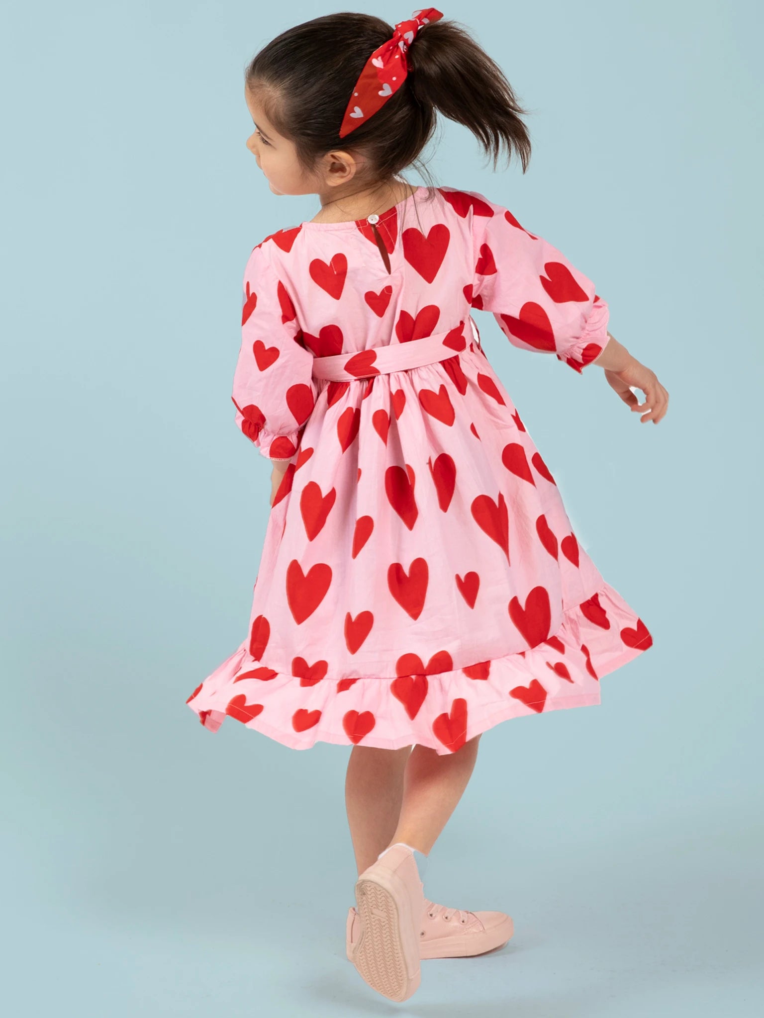 A young girl with a ponytail wears the Big Hearts Big Heart Girl's Midi Dress—handprinted cotton with red heart patterns—paired with pink sneakers and a matching red headband, set against a light blue background.