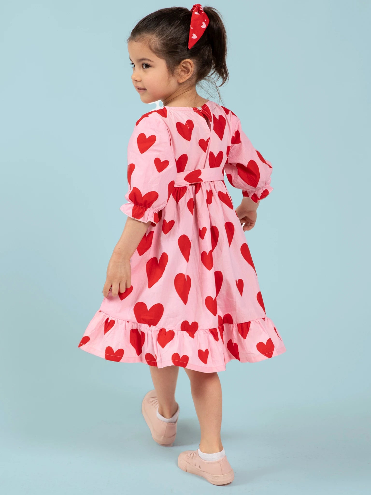 A young girl with a ponytail wears the Big Hearts Big Heart Girl's Midi Dress—handprinted cotton in pink with red hearts—and pink shoes, standing against a light blue background.
