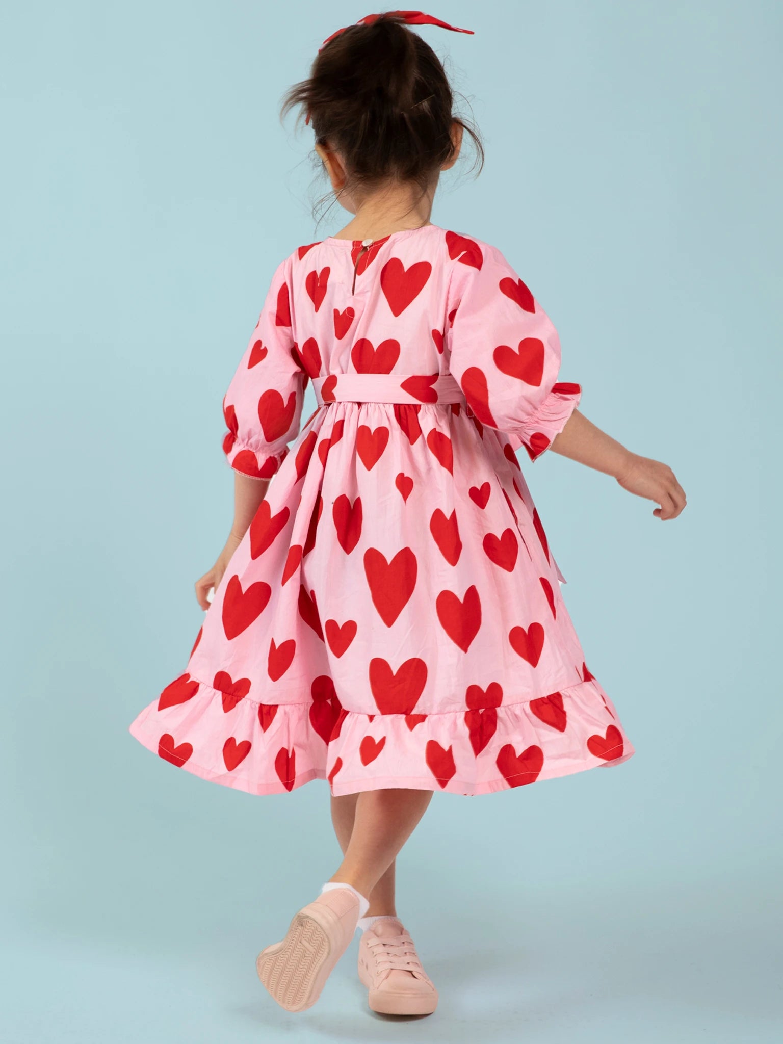A young girl with a red ribbon wears the Big Hearts Big Heart Girl's Midi Dress—ethically made in India from handprinted cotton with large red hearts—paired with pink shoes, standing against a light blue background, facing away.