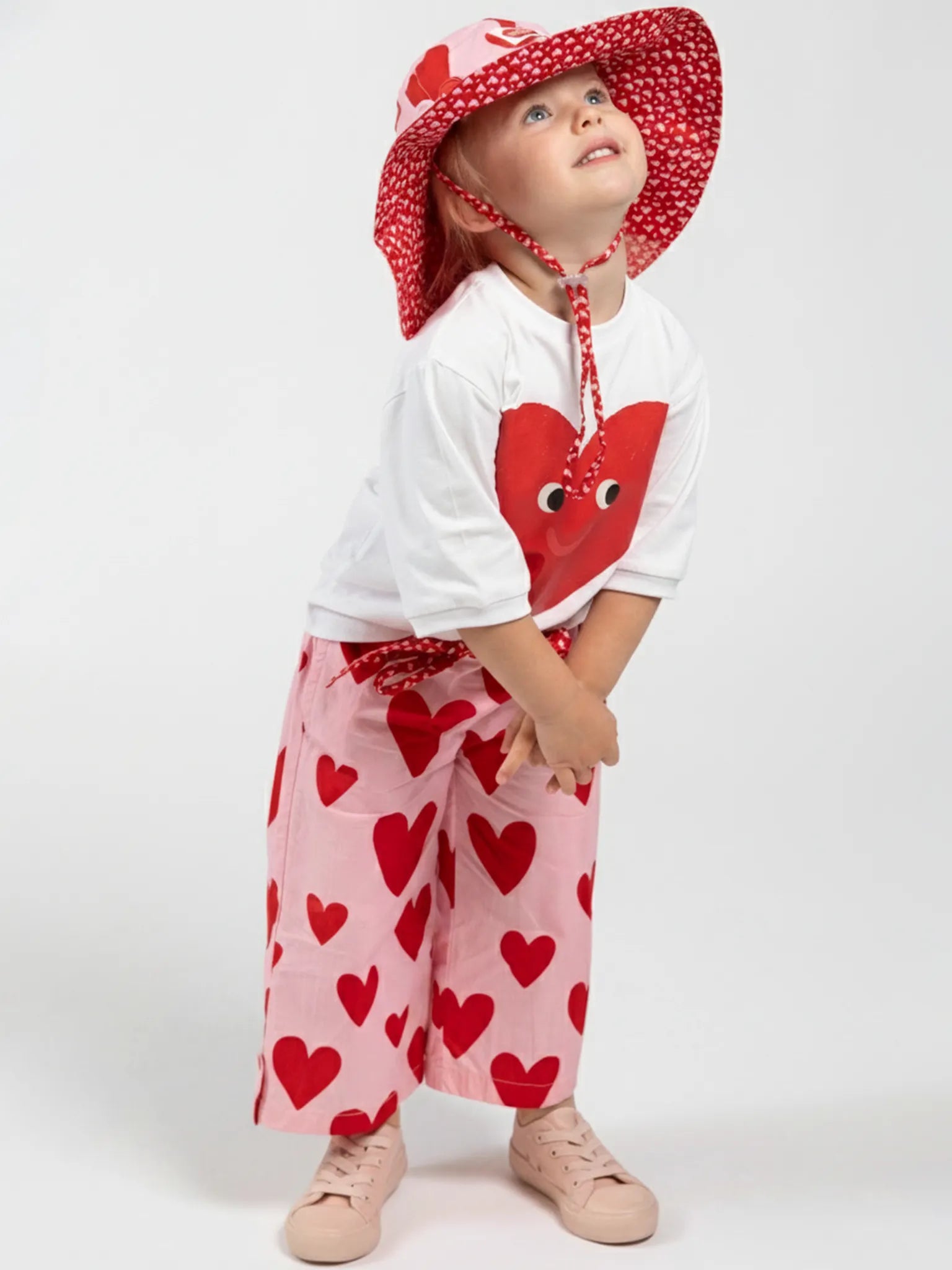 A young child in a white shirt with a red heart graphic, pink pants, and pink shoes wears the Big Hearts Big Heart Ponytail Hat with red hearts while standing against a plain white background.