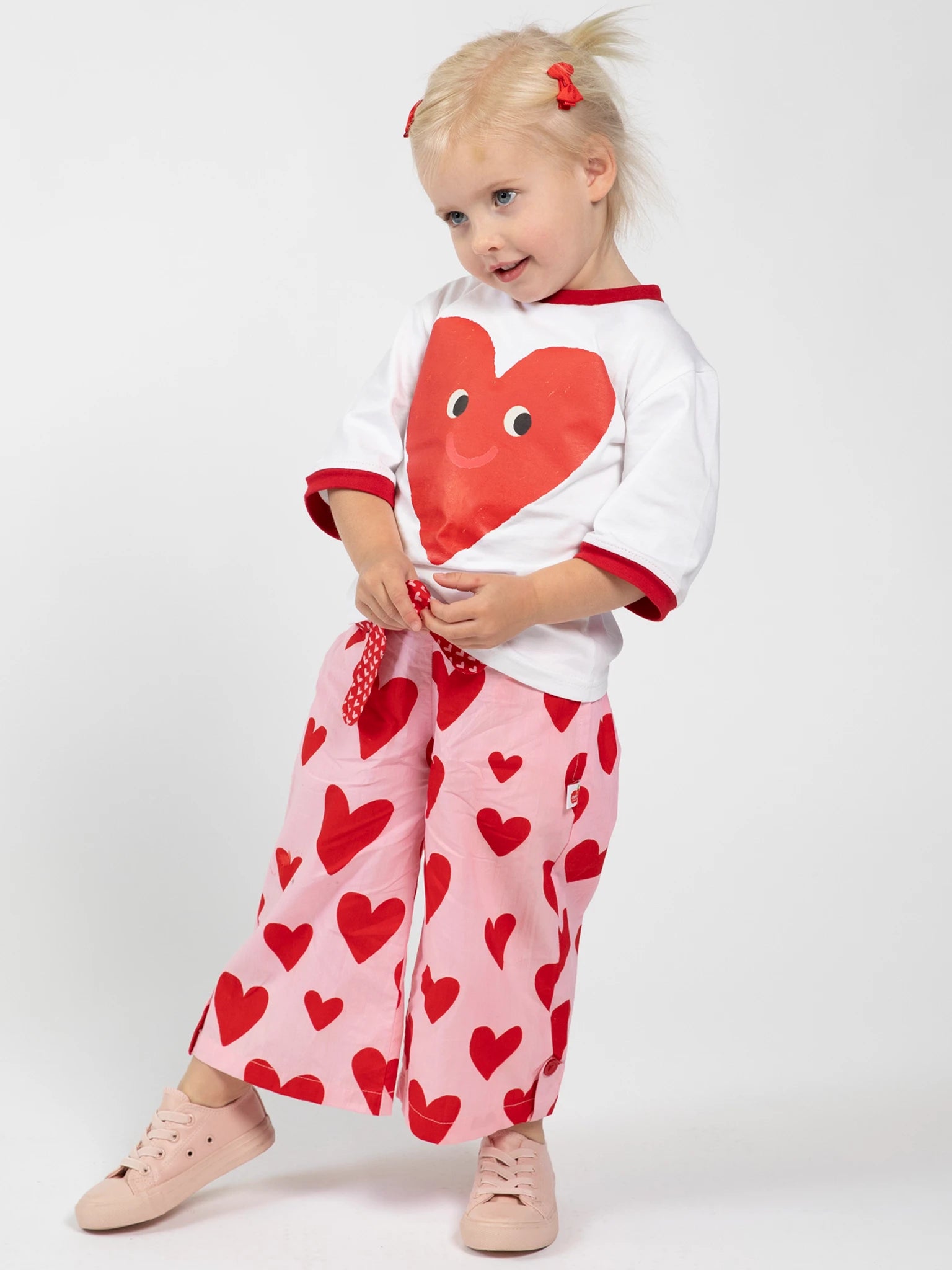 A young blonde girl stands against a plain white background, wearing Big Hearts’ Big Heart Wide-Leg Pants—elastic-waisted pink pants with red hearts—and a white shirt featuring a bold red heart graphic.