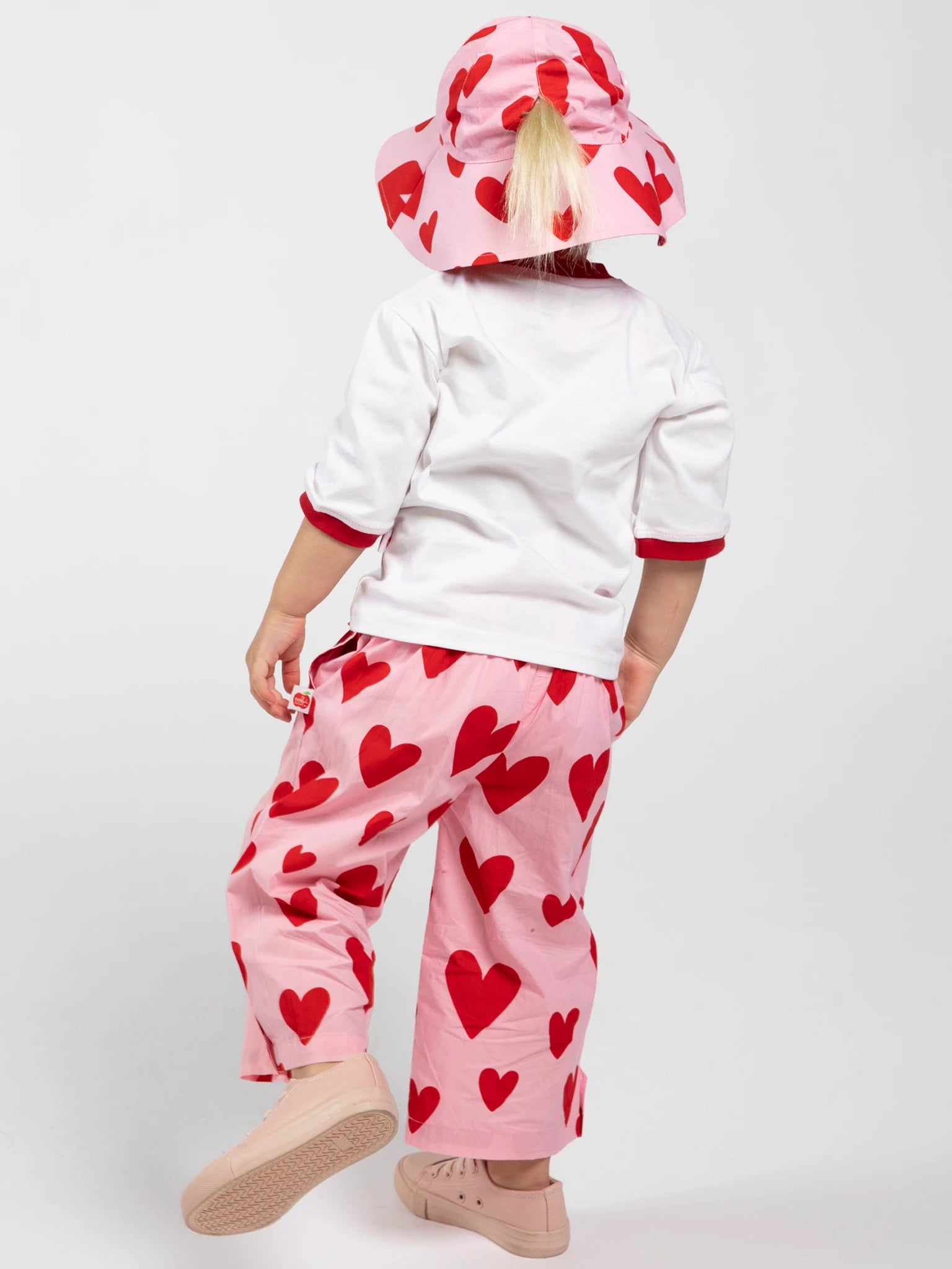 A child faces away, wearing a white shirt, pink pants, and a Big Hearts Big Heart Ponytail Hat—an adjustable kids hat with red heart patterns—against a plain white background.