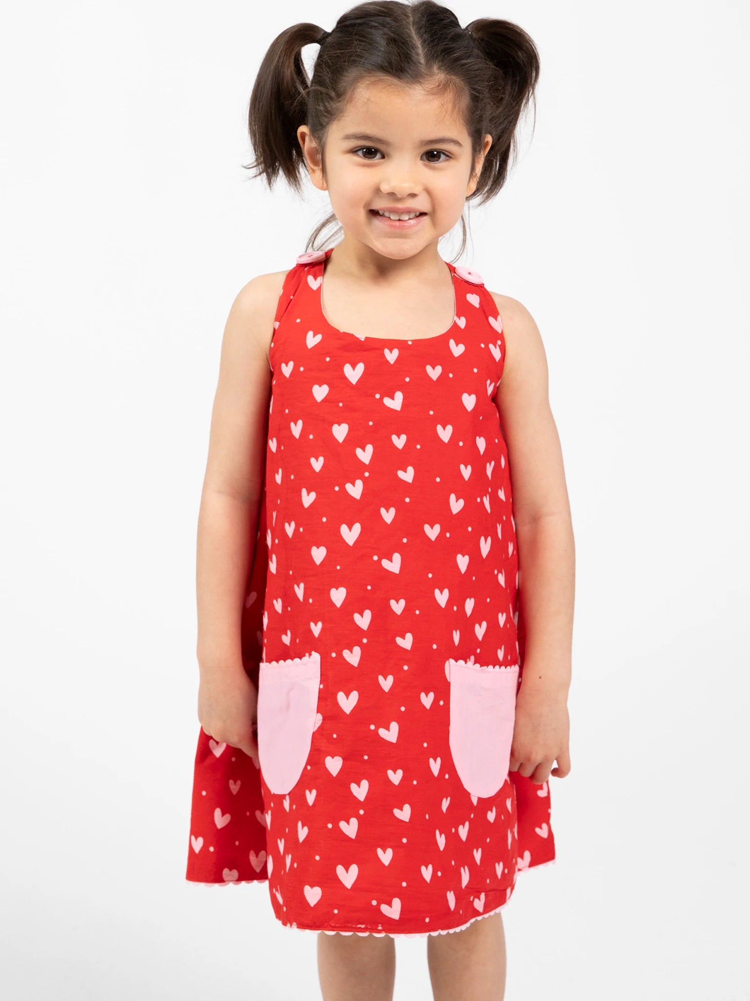 A young girl with pigtails wears the Big Hearts 'Big Heart Gracie Reversible Dress,' a red girls' cotton dress with white heart prints and two pink pockets, standing against a plain white background.