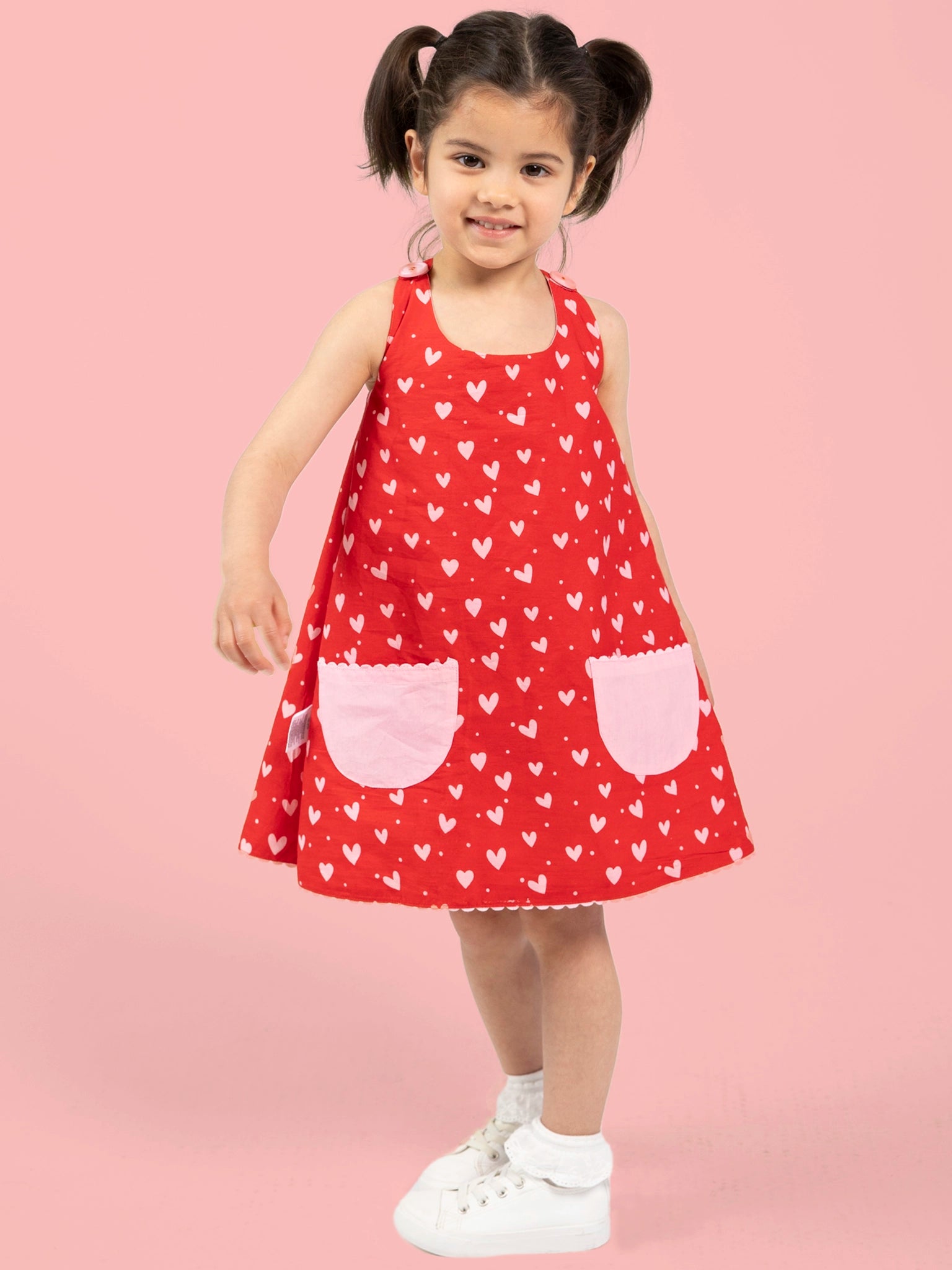 A young girl with pigtails wears the Big Hearts Big Heart Gracie Reversible Dress, featuring red fabric with white heart patterns and pink pockets, styled with white socks and sneakers, standing against a pink background.