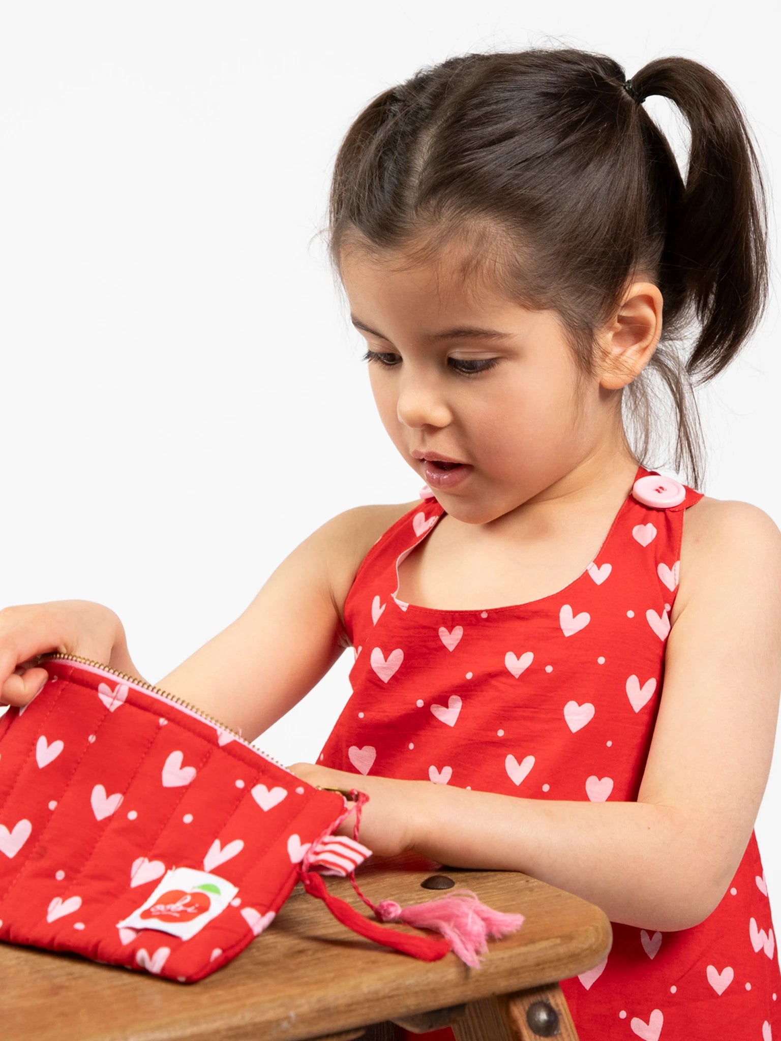 A young girl in a red heart-print dress sits at a wooden table, looking into the Oobi Heart Print Quilted Pouch with a gold zipper.