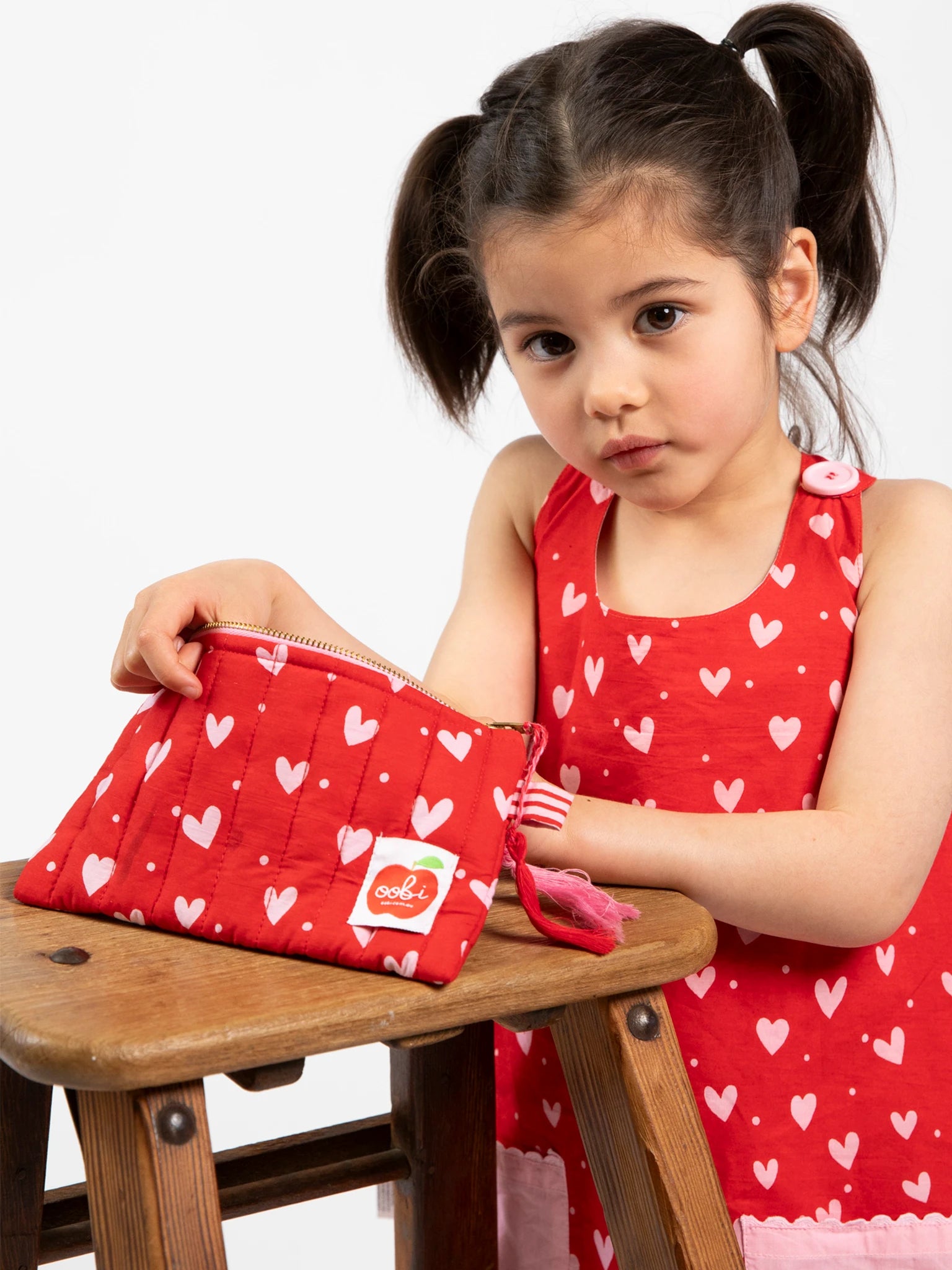 A young girl with dark pigtails sits on a wooden stool against a plain background, wearing a red dress with white hearts and holding the Oobi Heart Print Quilted Pouch.