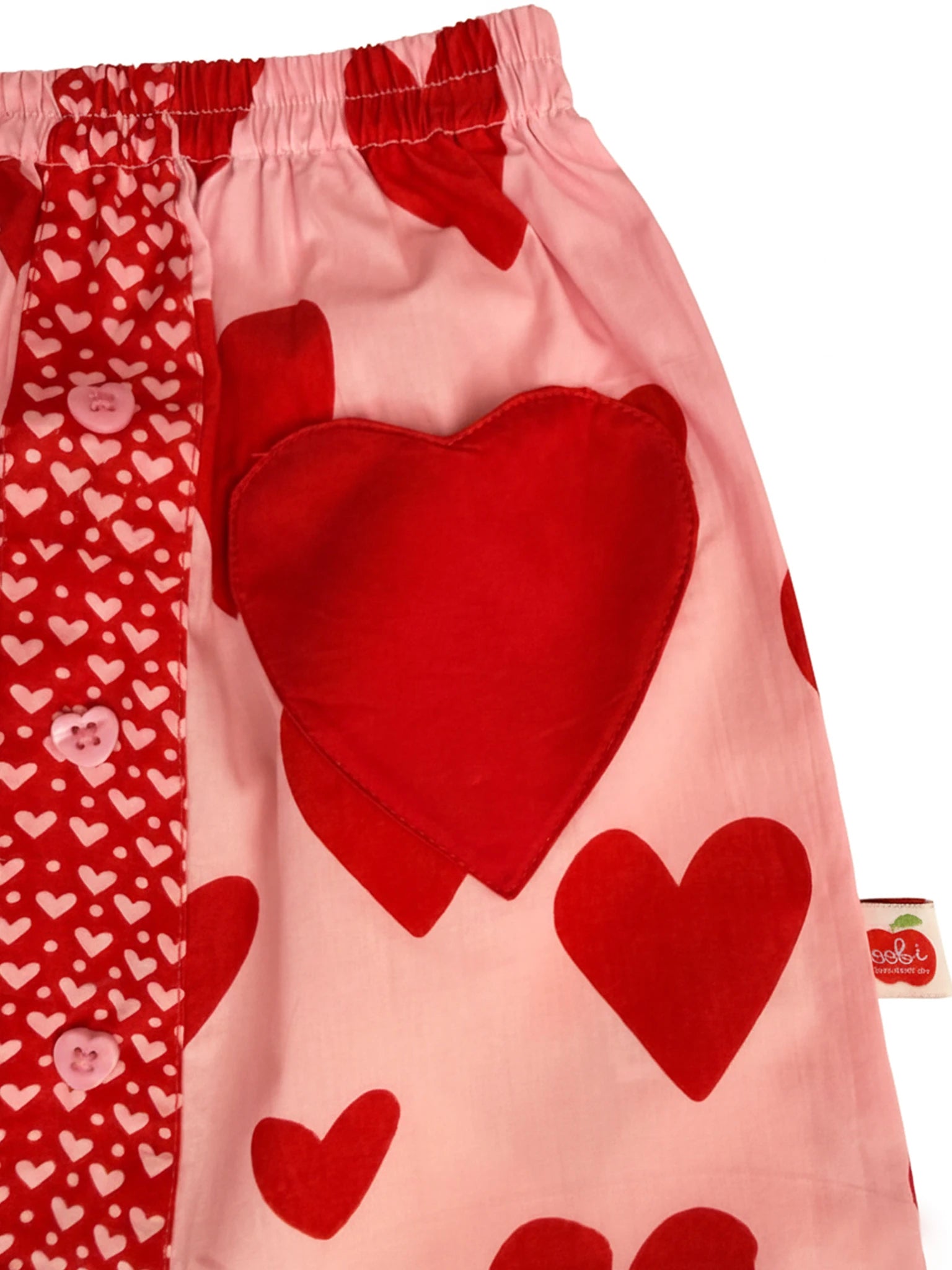 Close-up of the Big Hearts Big Heart Pocket Skirt in hand-printed cotton, featuring pink and red heart patterns, a large heart-shaped pocket, a fabric panel with small white hearts, and pink buttons.