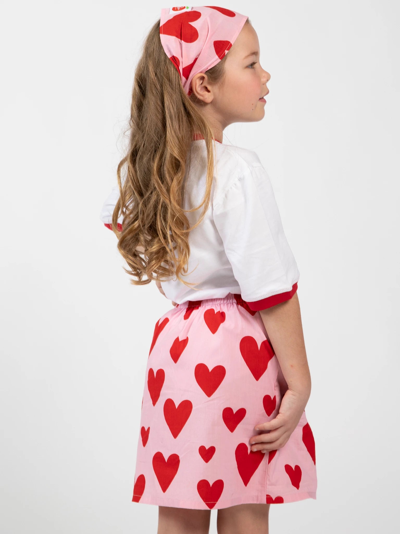 A young girl stands in profile wearing a white top, the Big Hearts Big Heart Pocket Skirt featuring hand-printed cotton with red heart patterns, and a matching headband.