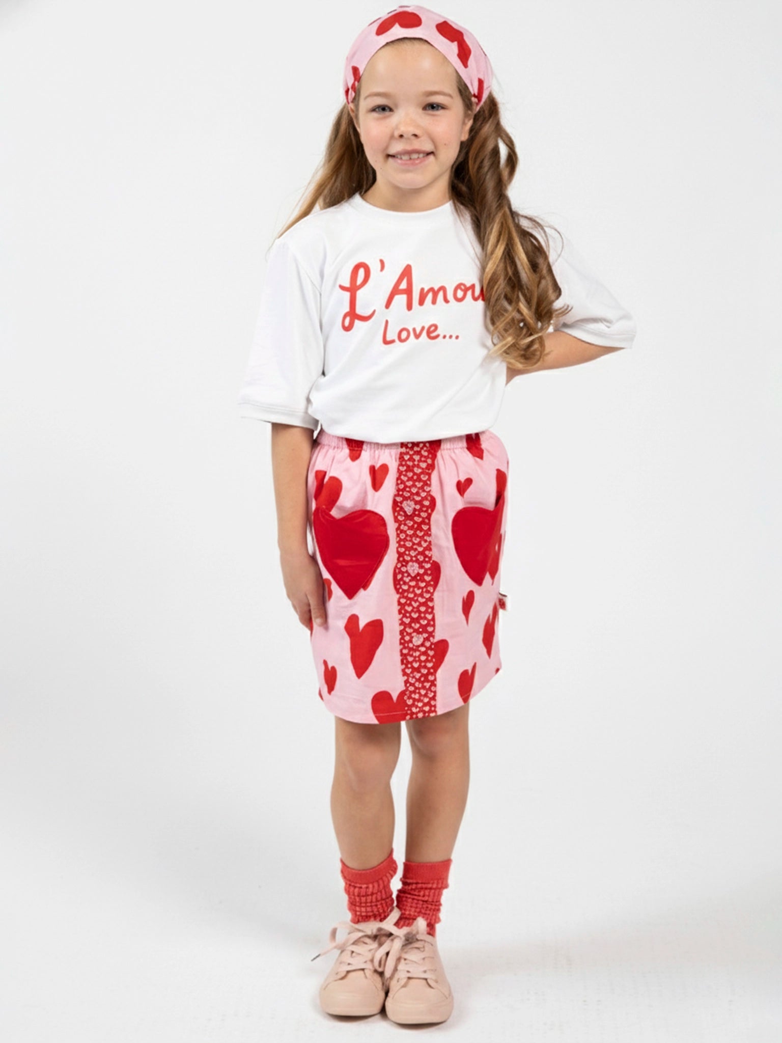 A young girl smiles in an oversized Essentials L’Amour (Love) Tee with Red Binding, a pink skirt with red hearts, matching headband, red socks, and pink shoes—all set against a white background.